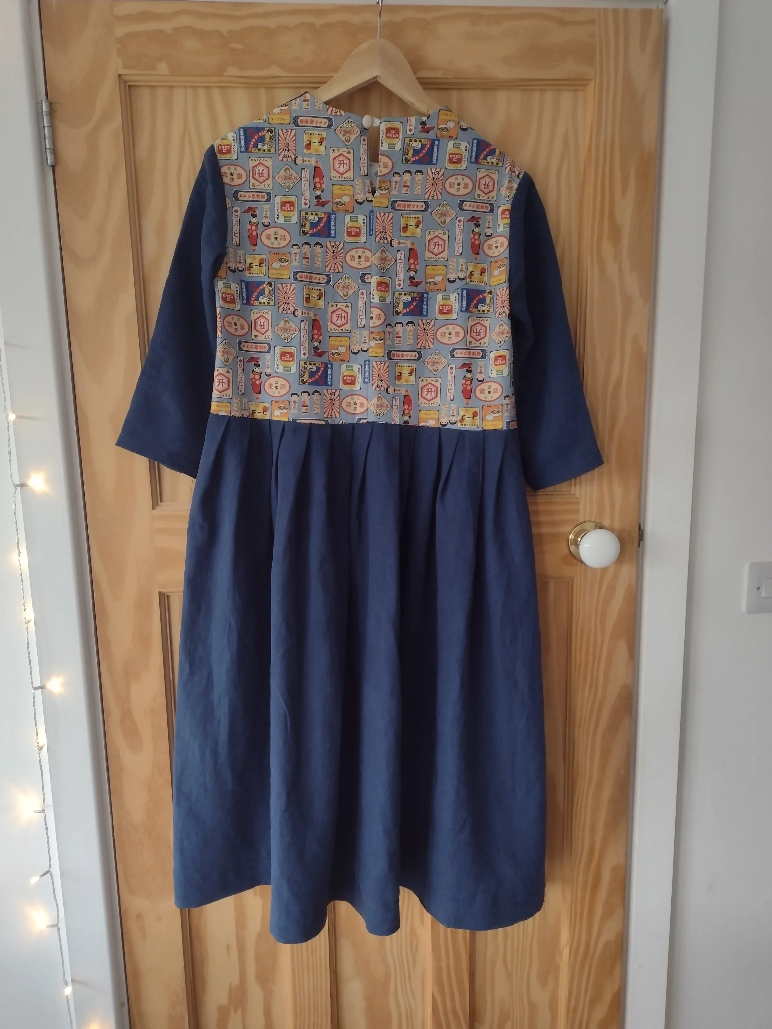 A dress hung on a wooden hanger on a wooden door. The dress has a top with colorful Japan-inspired patterns featuring various illustrations and symbols, and a long, dark blue skirt.
