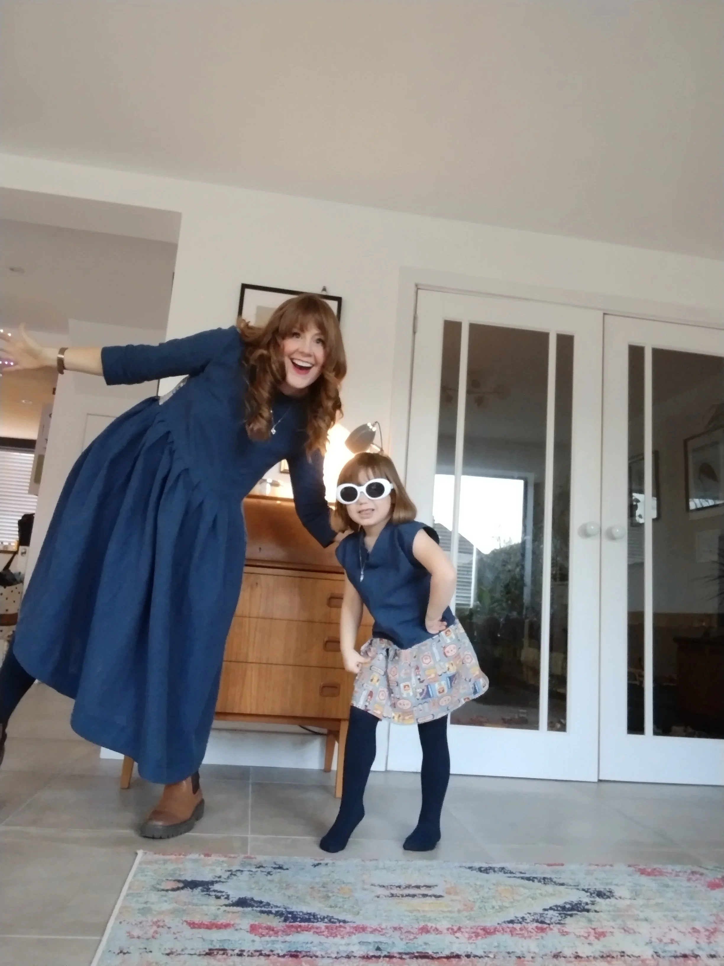 A woman and a young girl are indoors. The woman has long, wavy brown hair and is wearing a blue dress. The girl has shoulder-length brown hair, is wearing white sunglasses, a dark sleeveless top, a colorful skirt, and dark tights. They are both smiling, with the woman leaning forward and the girl striking a pose. Behind them are sliding glass doors and a wooden sideboard.