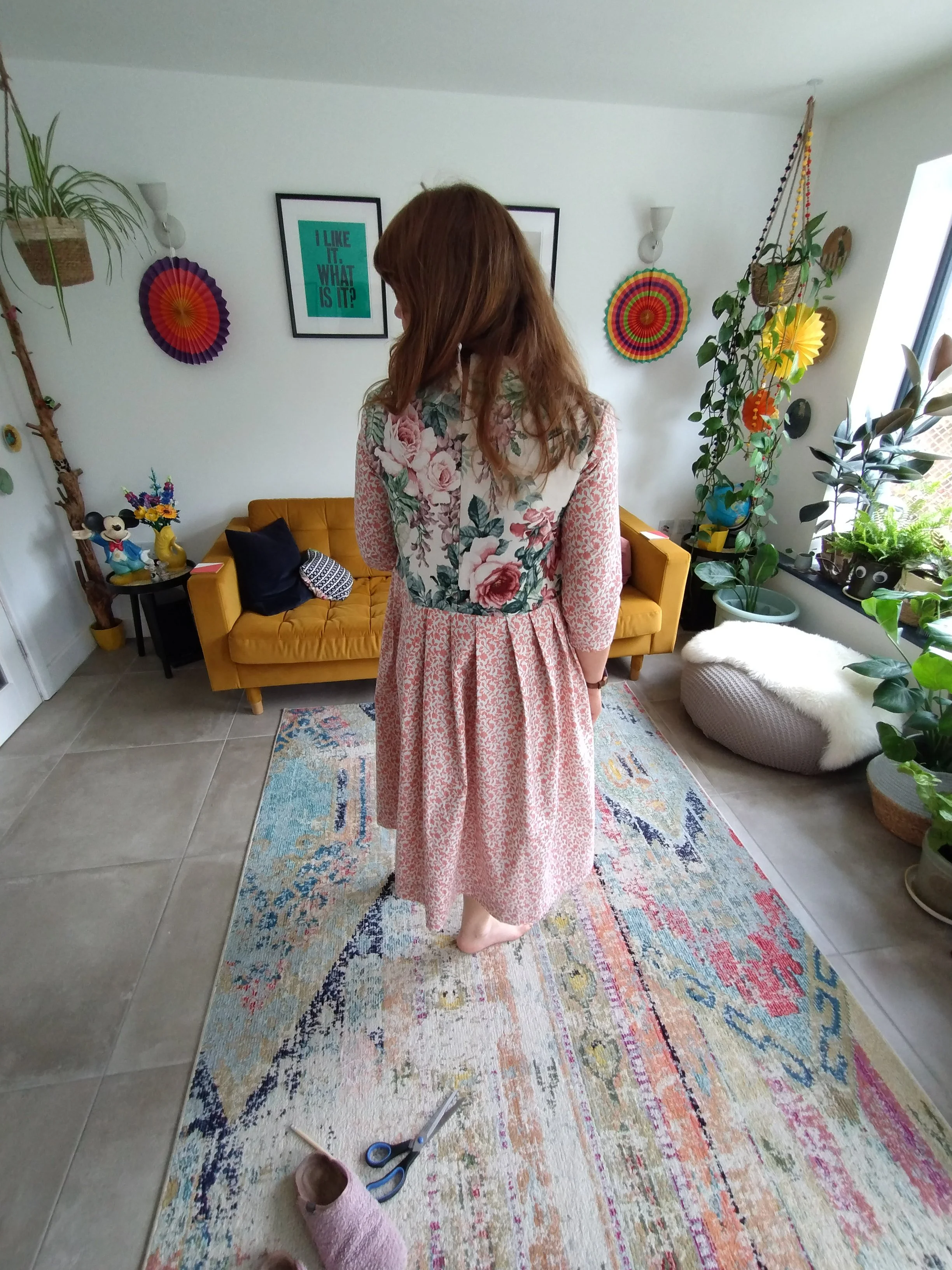 A woman in a long pink floral dress standing on a colorful rug in a living room filled with houseplants and decorations.