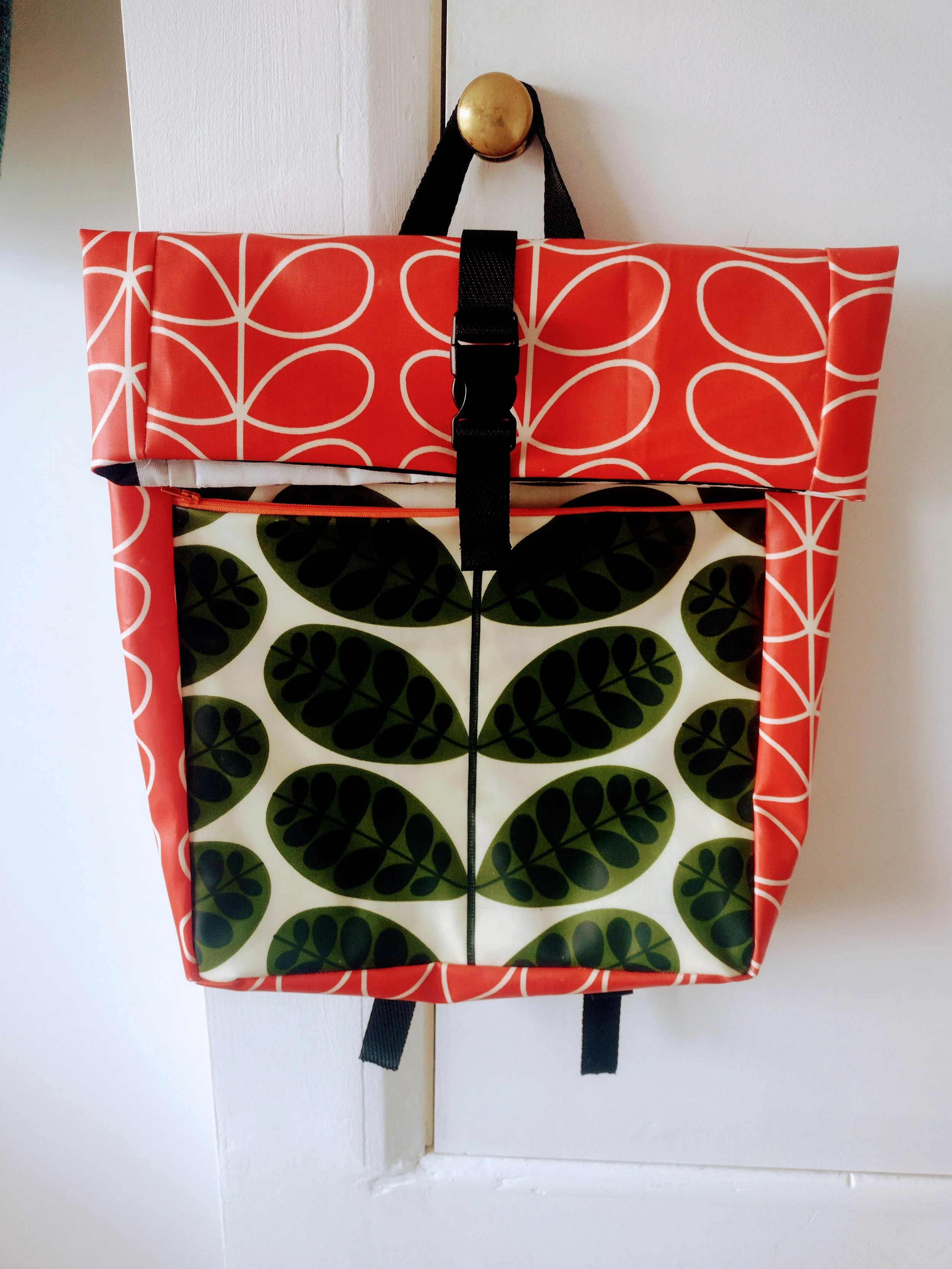 A colorful fabric bag hanging from a brass doorknob. The bag has a red top with white interlocking circles, and a white bottom with a green leafy pattern.