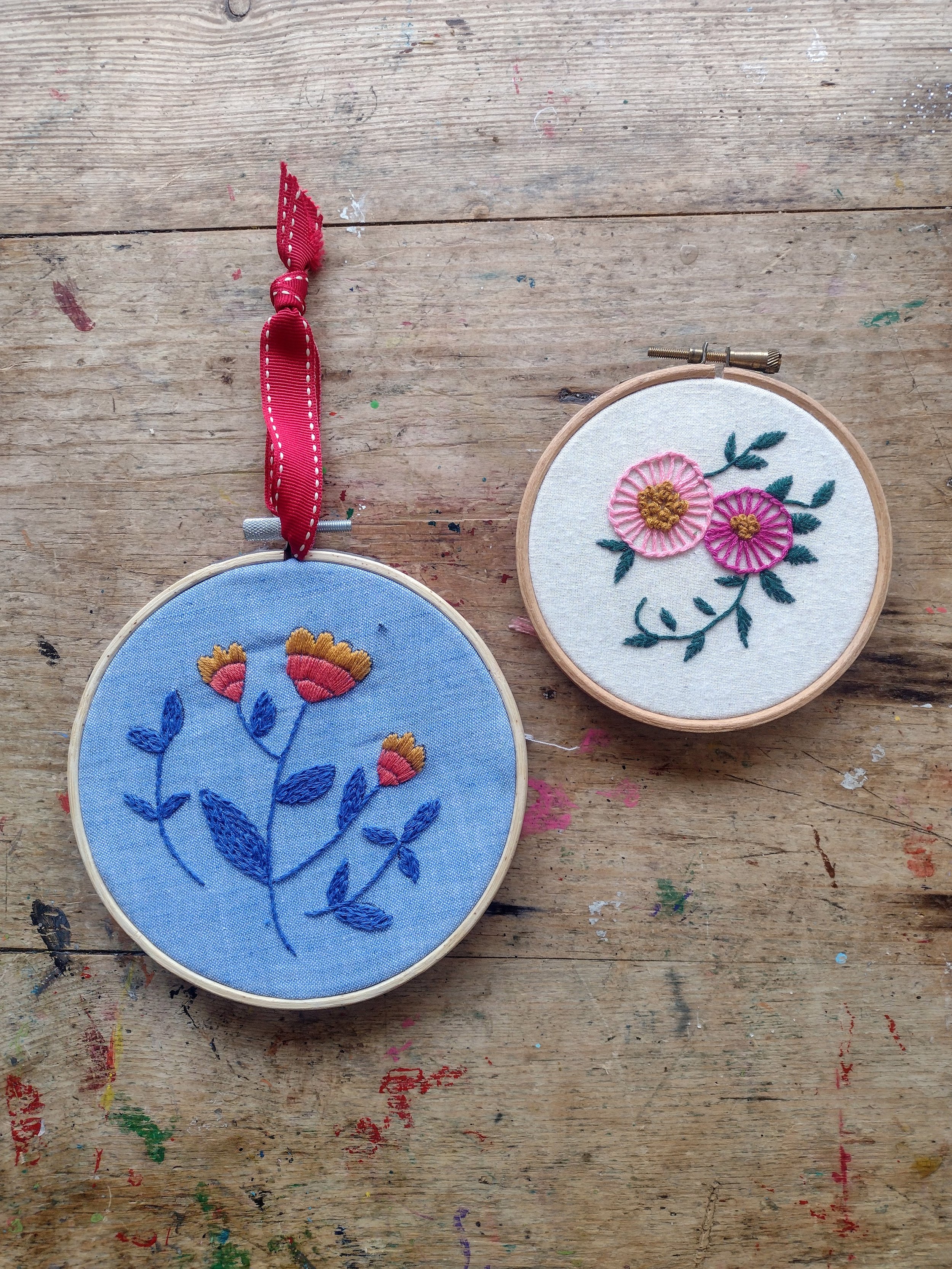 Two embroidery hoops with floral designs on a wooden table. The hoop on the left has a blue fabric with dark blue and pink flowers. The hoop on the right has a white fabric with pink and yellow flowers and green leaves.