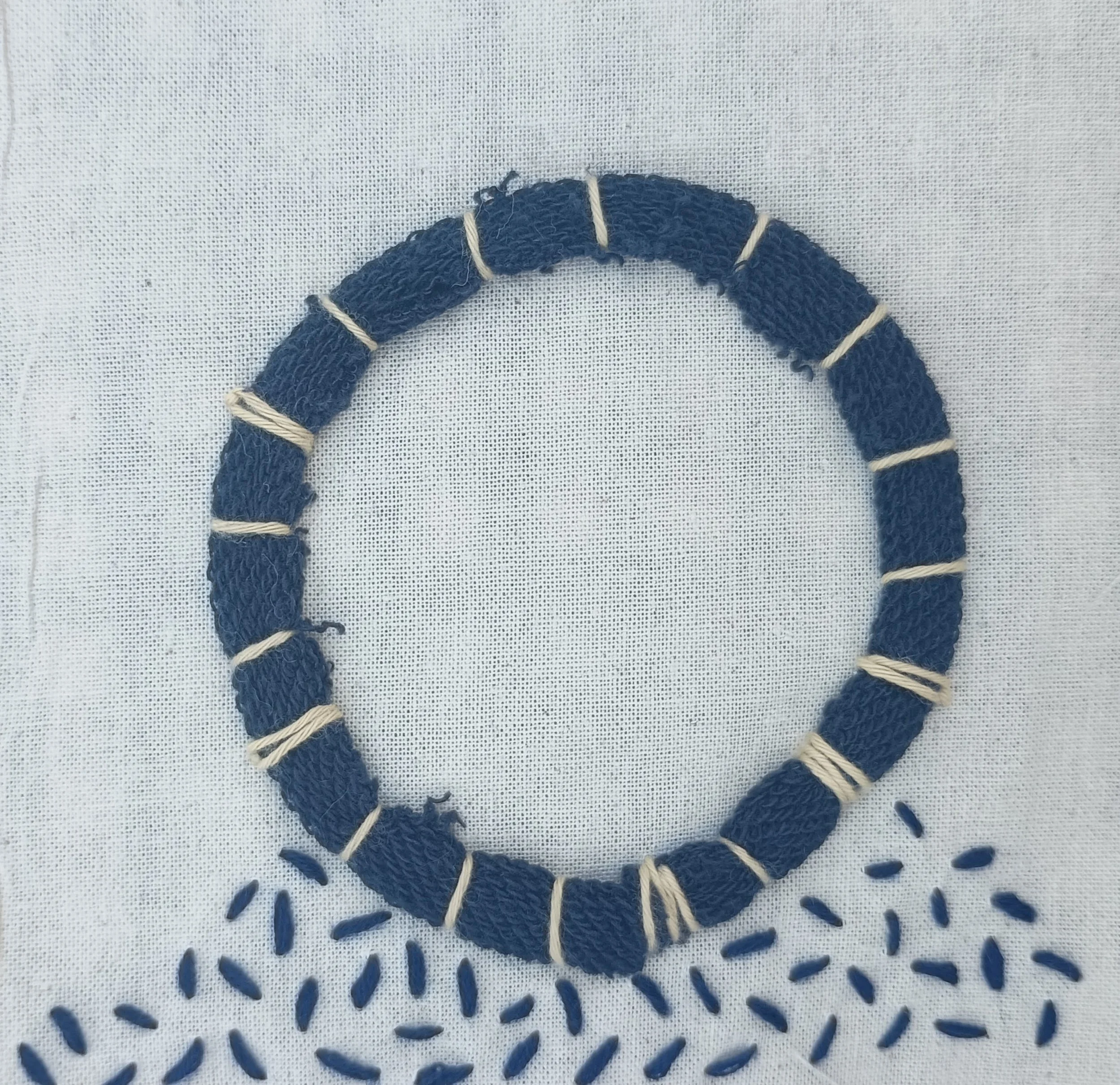 A close-up of a dark blue fabric or thread coiled into a circle, with white or cream-colored thread wrapped around parts of it, placed on a white textured fabric background.