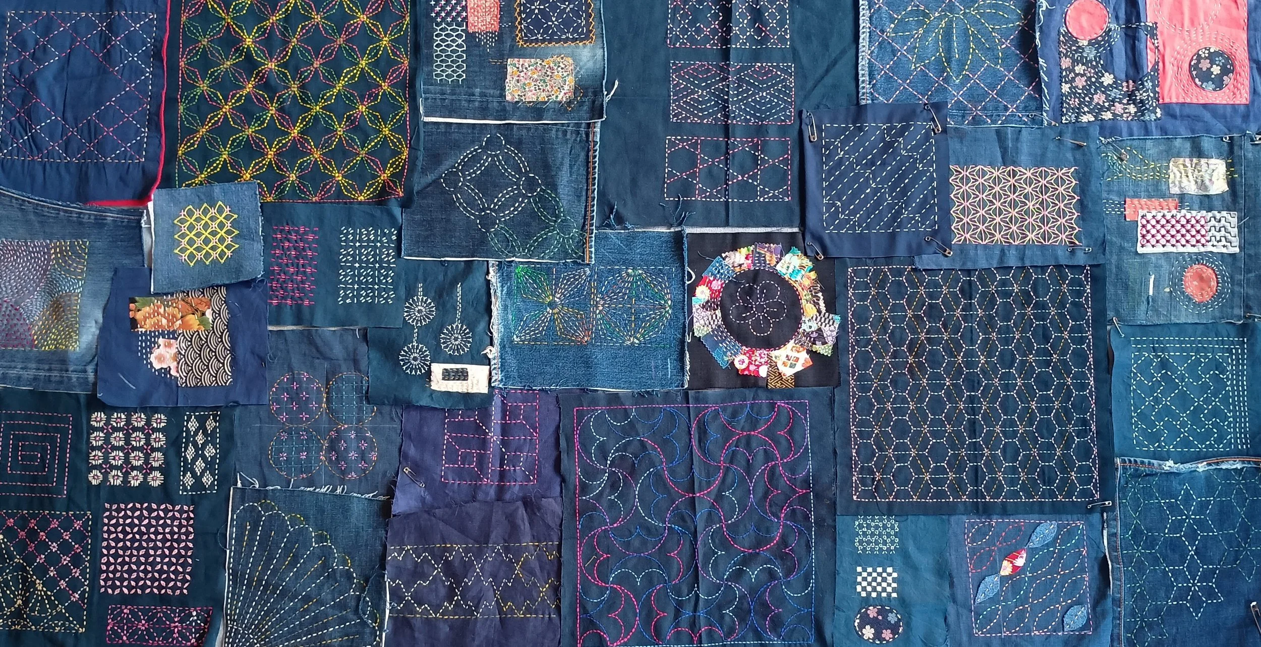 A large wall display of various pieces of fabric with intricate embroidery and quilting, mostly featuring geometric patterns and vibrant thread colors, on a dark blue background.
