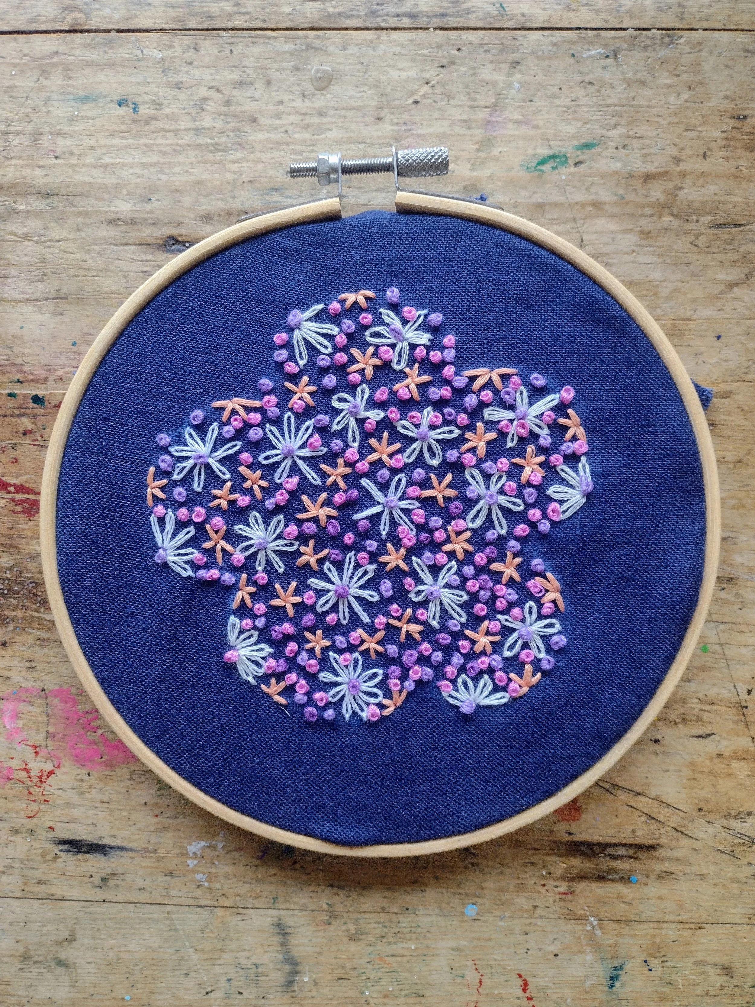 A close-up of an embroidery hoop with purple, white, and peach floral embroidery on dark blue fabric, placed on a wooden surface.