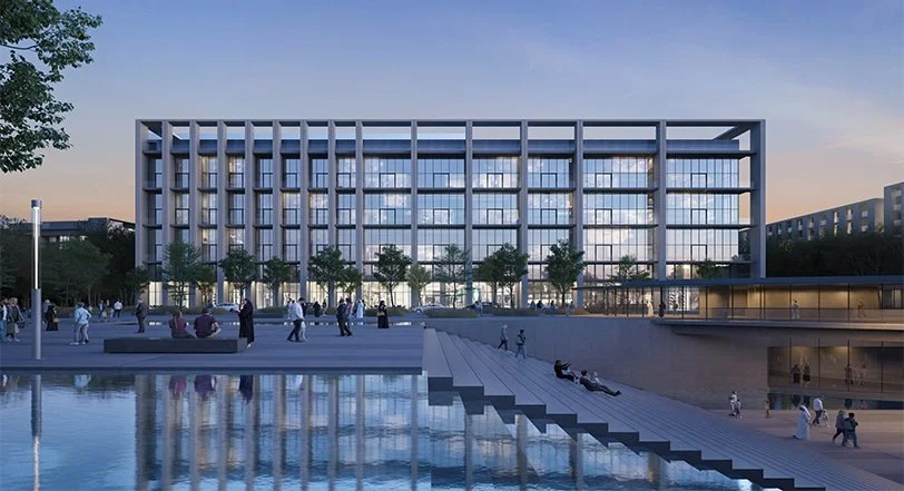 Modern multi-story building with glass windows, reflecting the cloudy sky, situated beside a body of water with stairs leading down to it. People are walking and sitting around the area, and trees line the foreground.