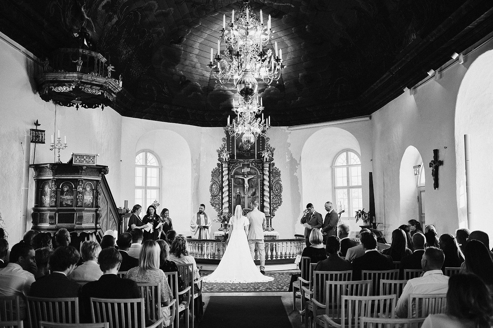 En svartvit bild av ett bröllop i en kyrka, med en brud och en brudgummasceremoni i fokus. Kyrkan har stora fönster, en kristusbild i centrum och kristallkronor i taket. Människor är samlade och sitta i trästolar och tittar på ceremonin.