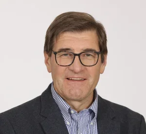 A middle-aged man with brown hair, glasses, wearing a dark suit and a blue checkered shirt, smiling against a plain white background.