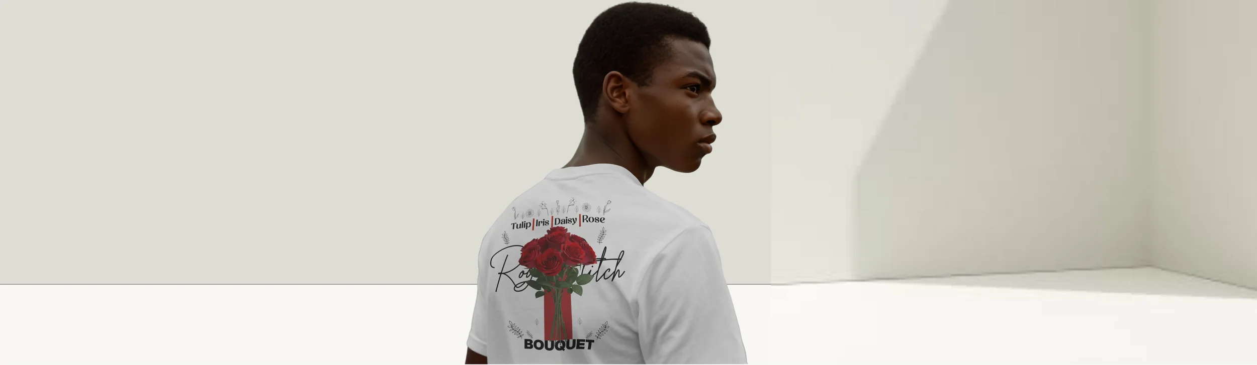 A young man with dark skin and short black hair stands against a plain beige wall, wearing a white T-shirt with a floral graphic and text related to roses.