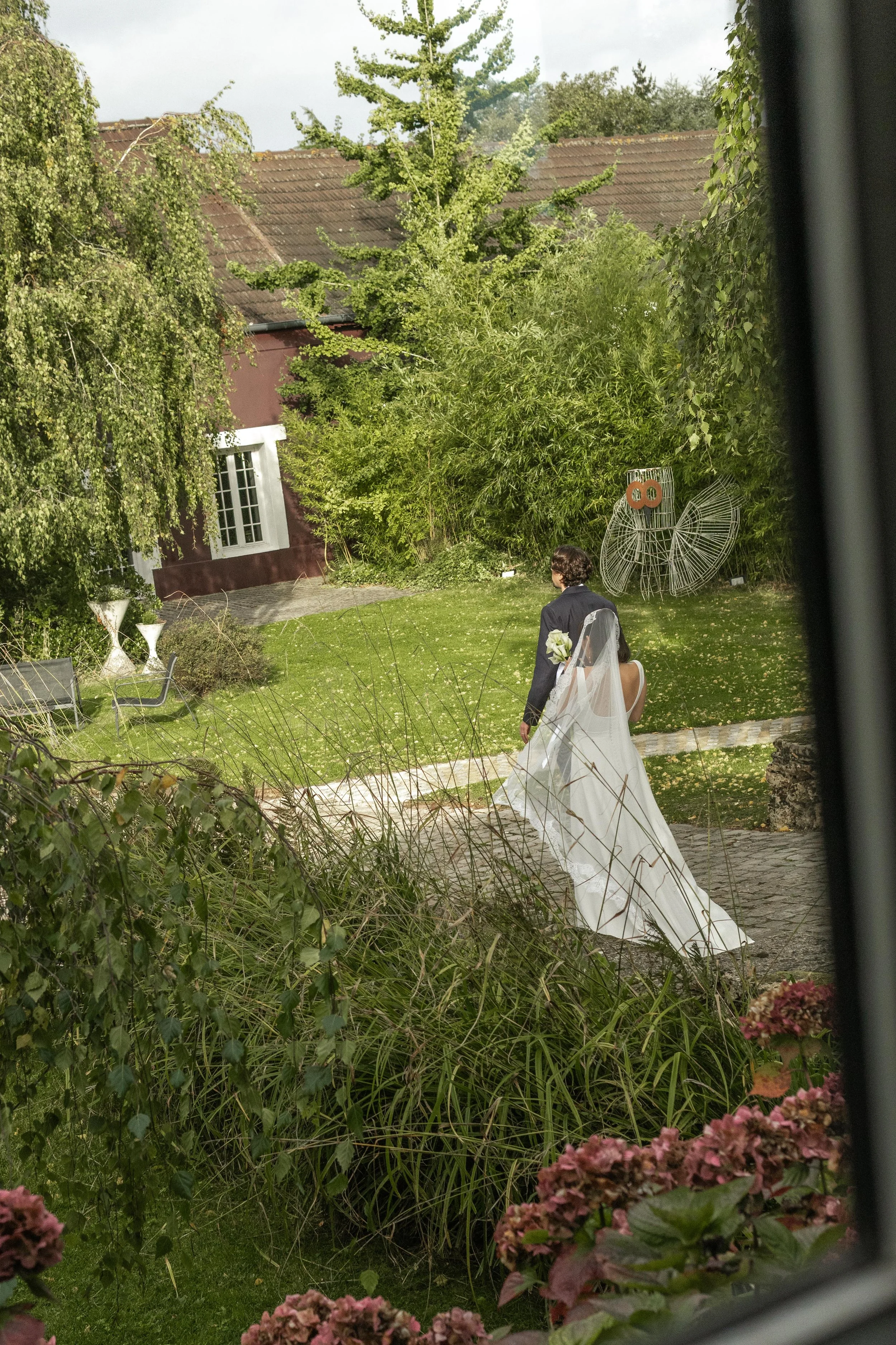 Un couple de mariés dans un jardin verdoyant, photographié depuis une fenêtre, avec des fleurs en premier plan.