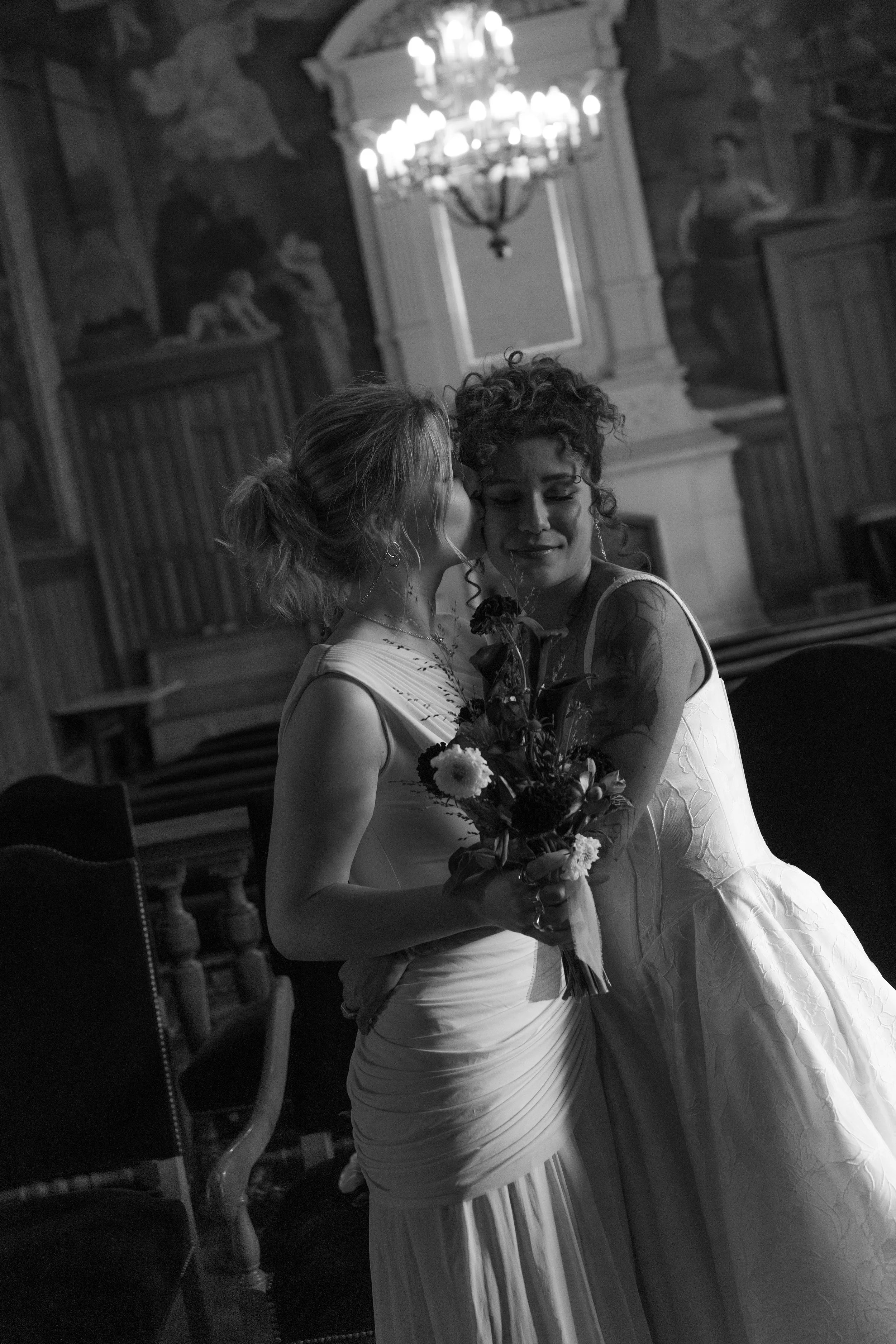 Deux femmes en robes de mariage s'embrassent dans une pièce ornée de peintures et d'un chandelier chandelier.