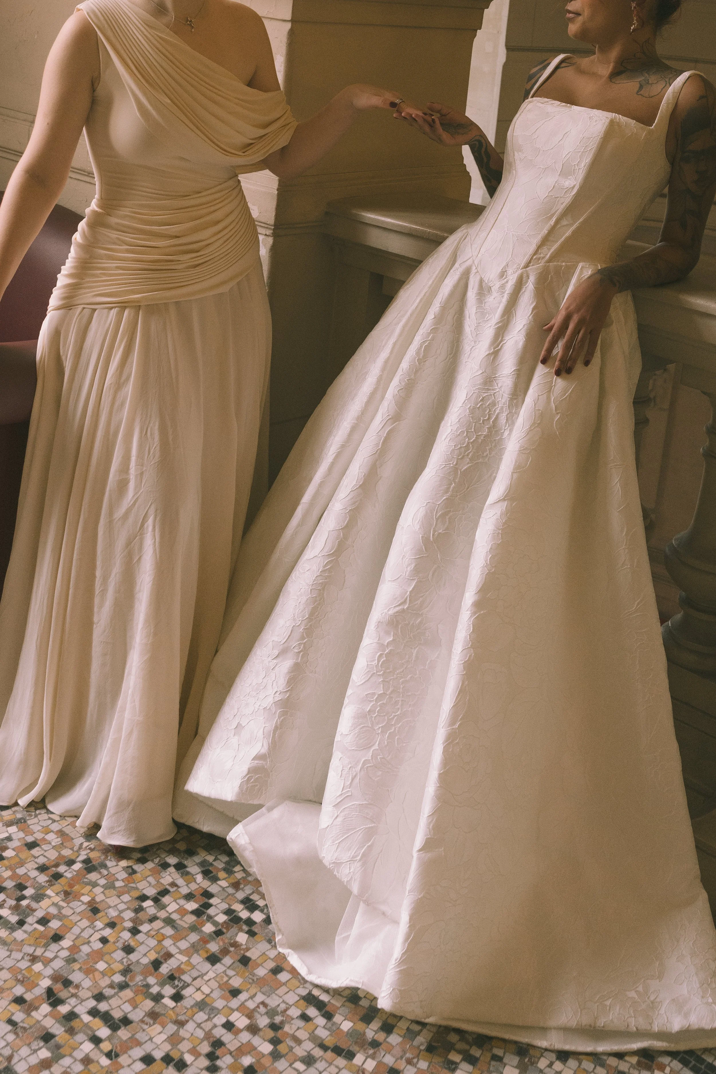 Deux femmes, dont l'une porte une robe de mariée blanche avec un corsage en satin et une jupe volumineuse, et l'autre porte une robe longue beige à un épaule, se tiennent dans un intérieur élégant et ancien.