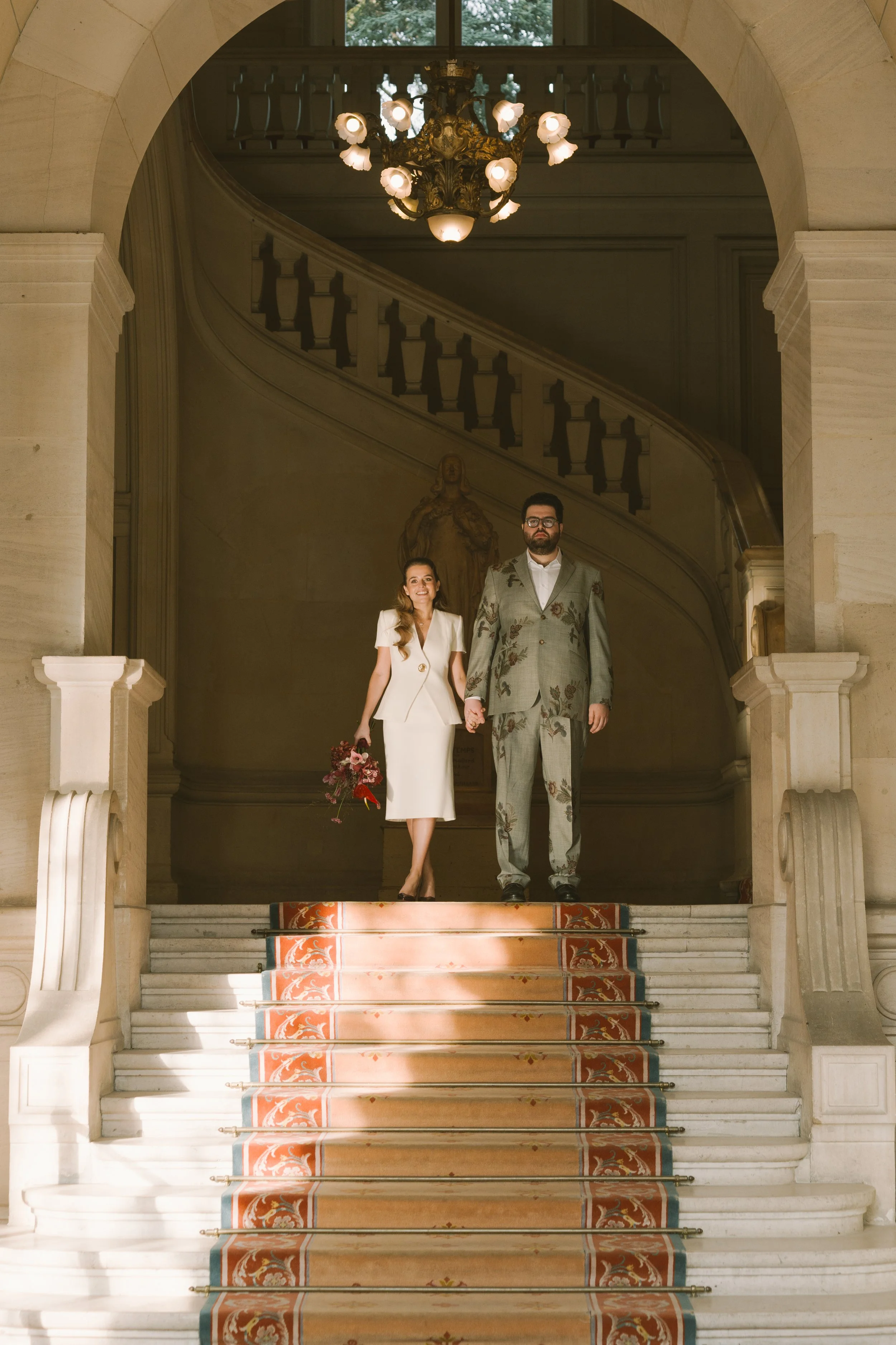 Un couple habillé élégamment, tenant la main, descend un escalier en marbre dans un bâtiment historique, avec une lampe suspendue au plafond et une sculpture en arrière-plan.