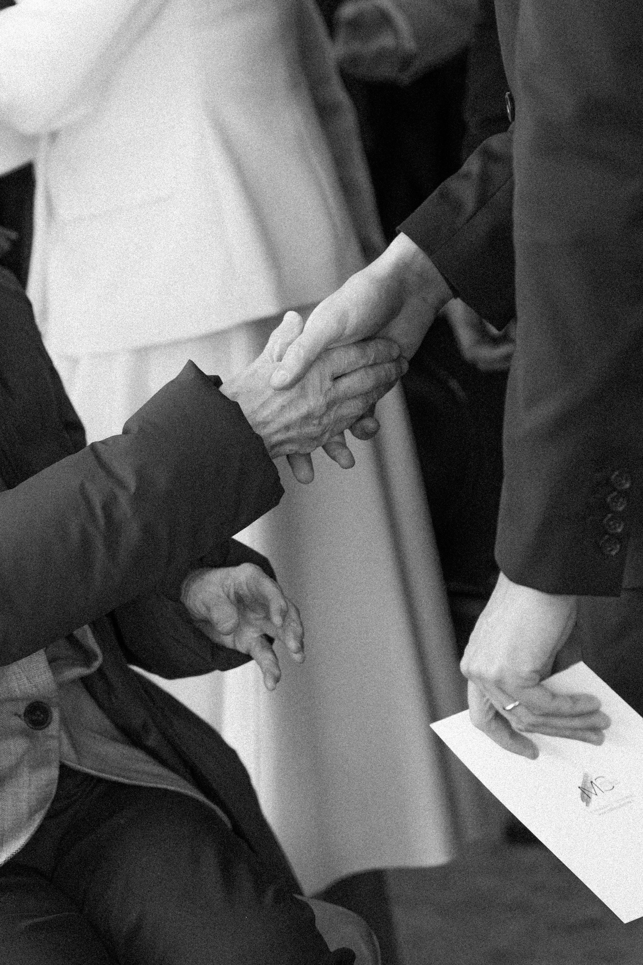 Deux personnes se serrent la main dans un contexte formel, une personne est assise avec une main pliée sur la jambe, une autre est debout, tenant une lettre ou un document.