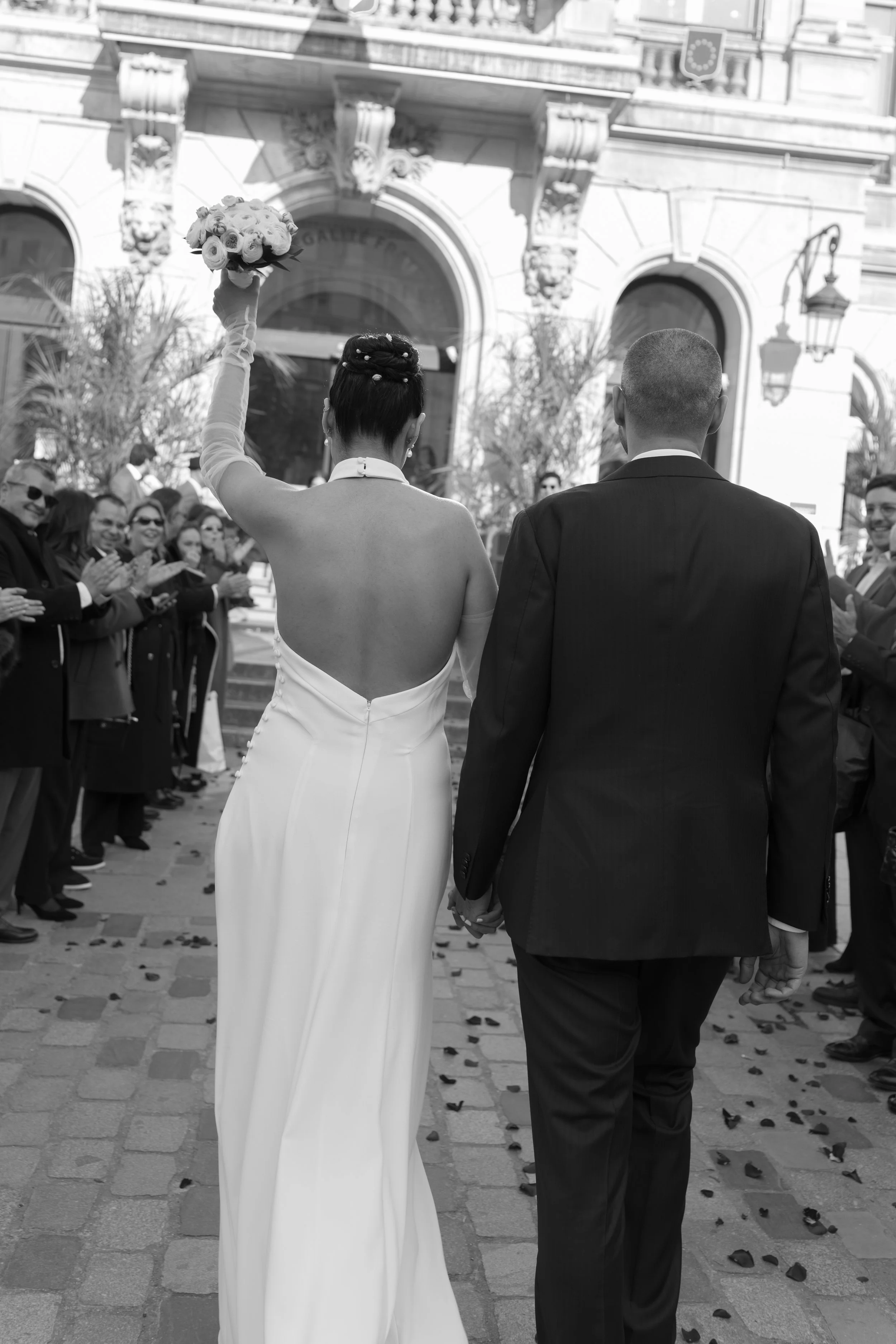 Un couple de mariés marchant main dans la main lors de leur mariage, la mariée porte une robe blanche longue et tient un bouquet de fleurs en l'air, avec une foule de personnes applaudi devant un bâtiment historique.