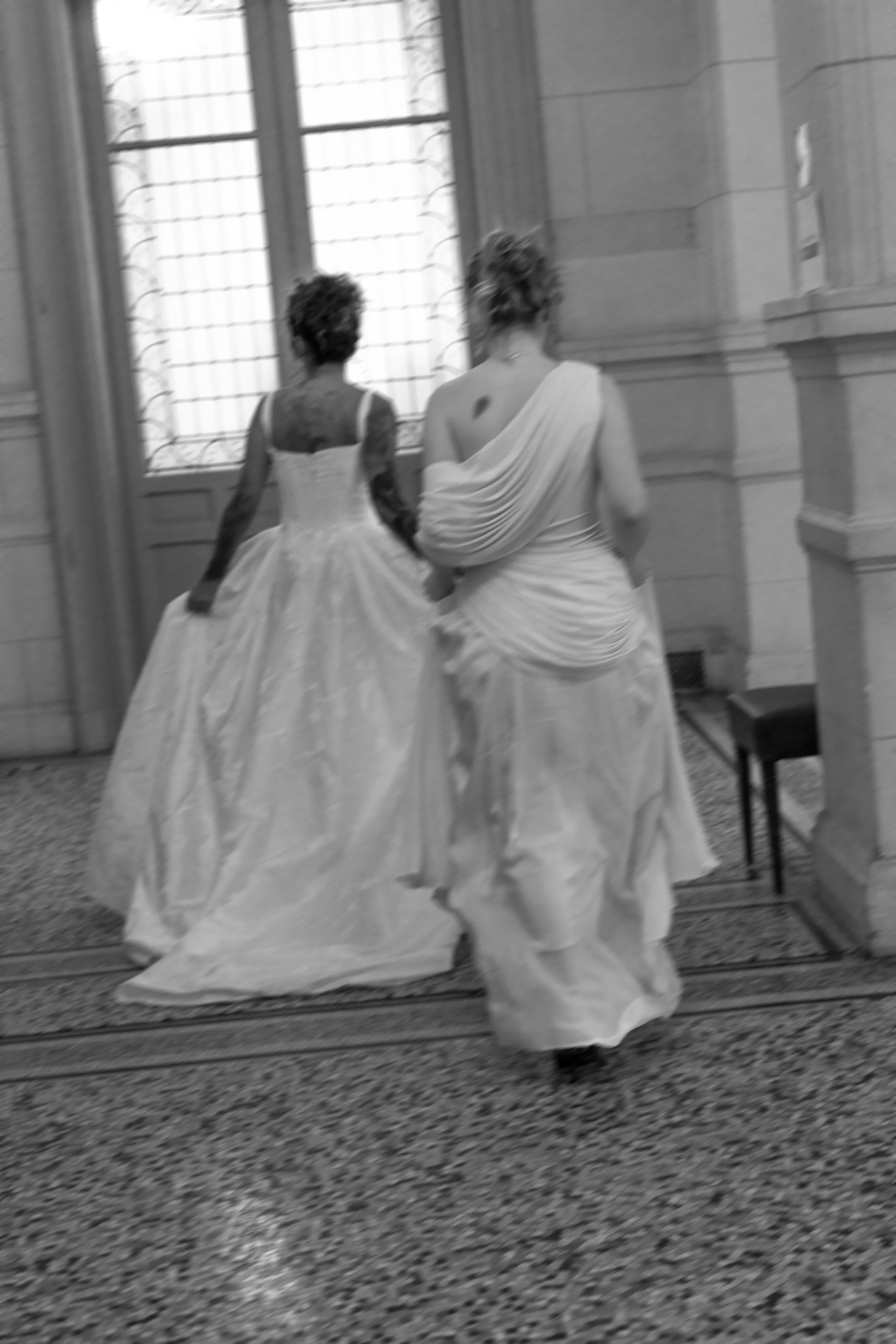 Deux femmes en robes de mariée marchent dans une salle avec des fenêtres et des murs en pierre.