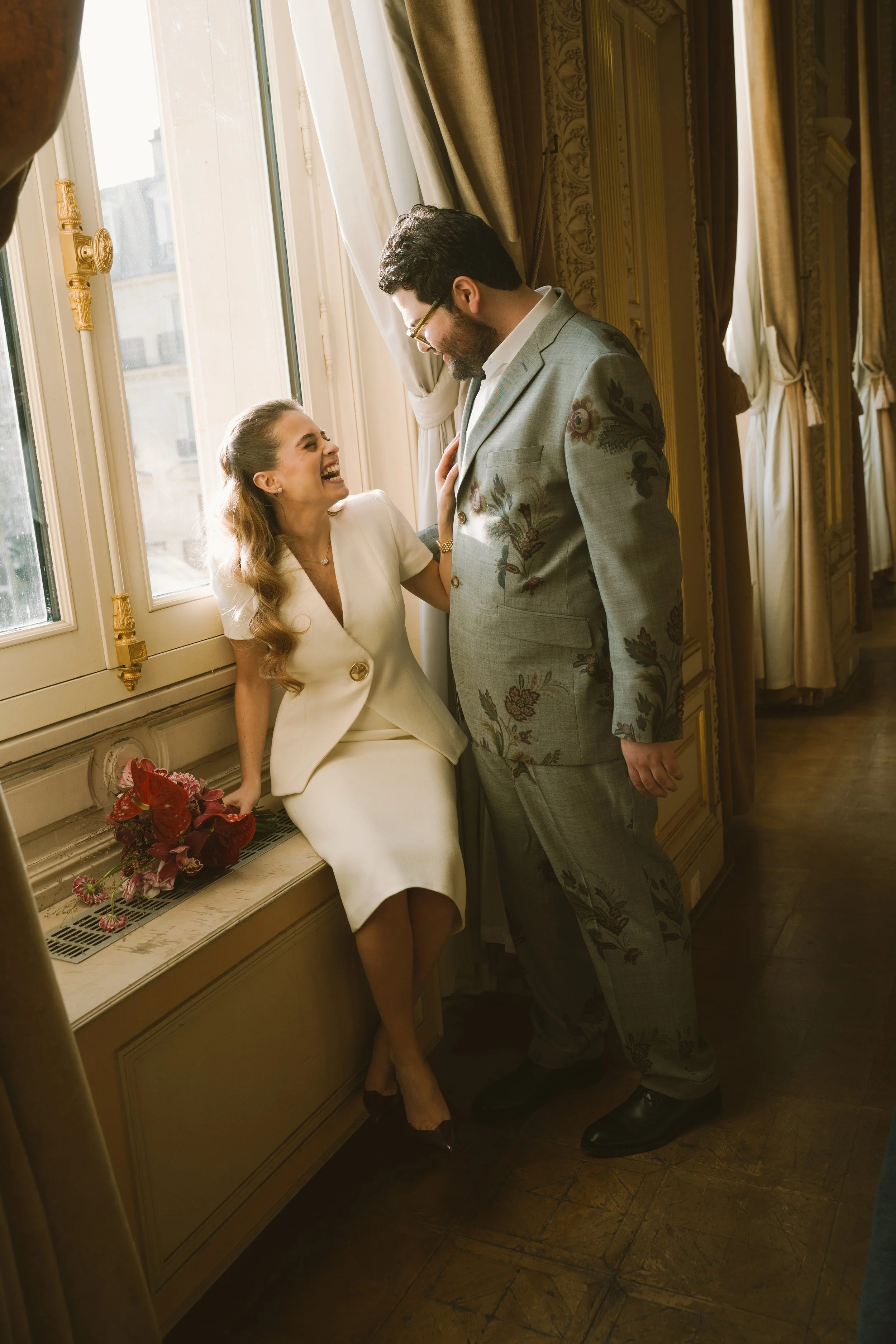 Un homme et une femme souriants, dans un intérieur élégant avec des rideaux en tissu doré, s'adonnent à une conversation joyeuse, la femme est assise sur une fenêtre avec un bouquet de fleurs à ses côtés.