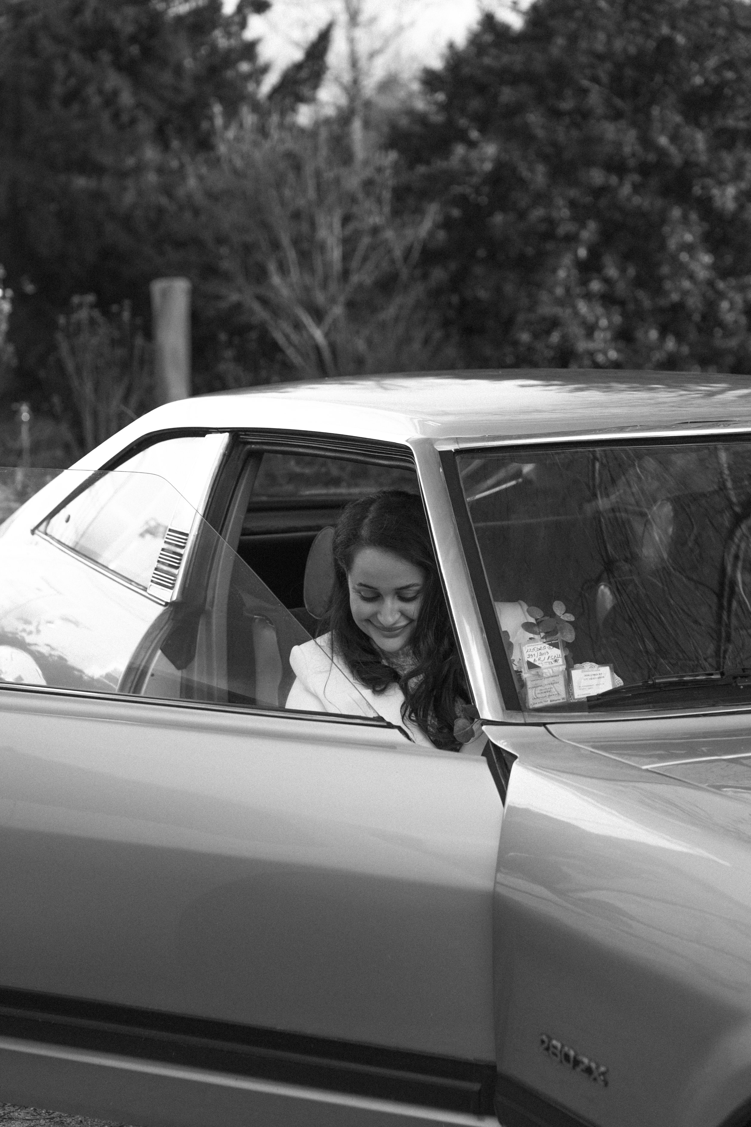 Femme souriante assise dans une voiture vintage, vue de face, avec un fond de nature arbres et ciel.