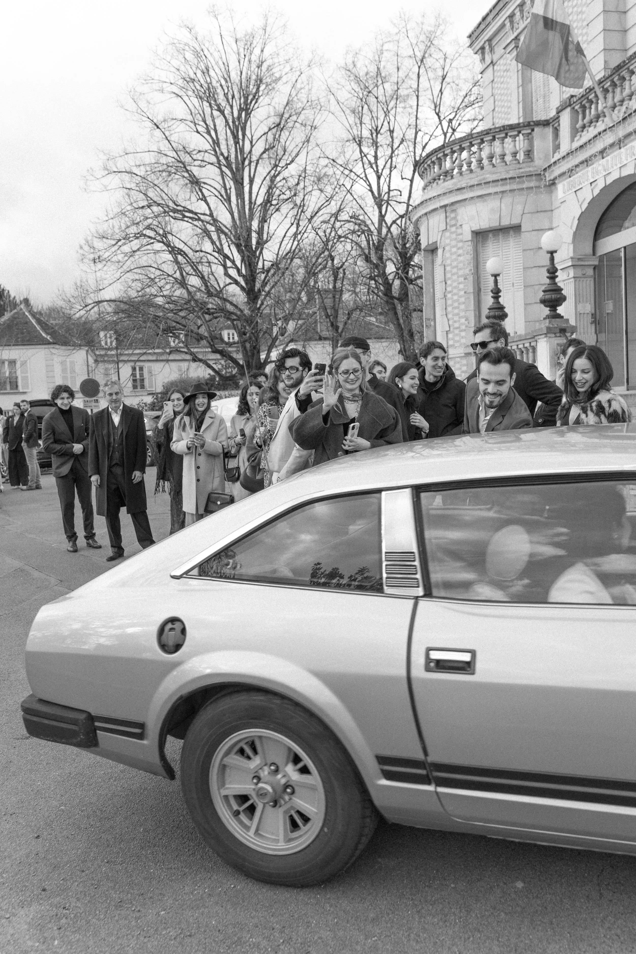 Une foule de personnes élégantes souriantes, certaines prenant des photos d'une voiture vintage qui passe devant une grande maison avec drapeau français, en hiver.