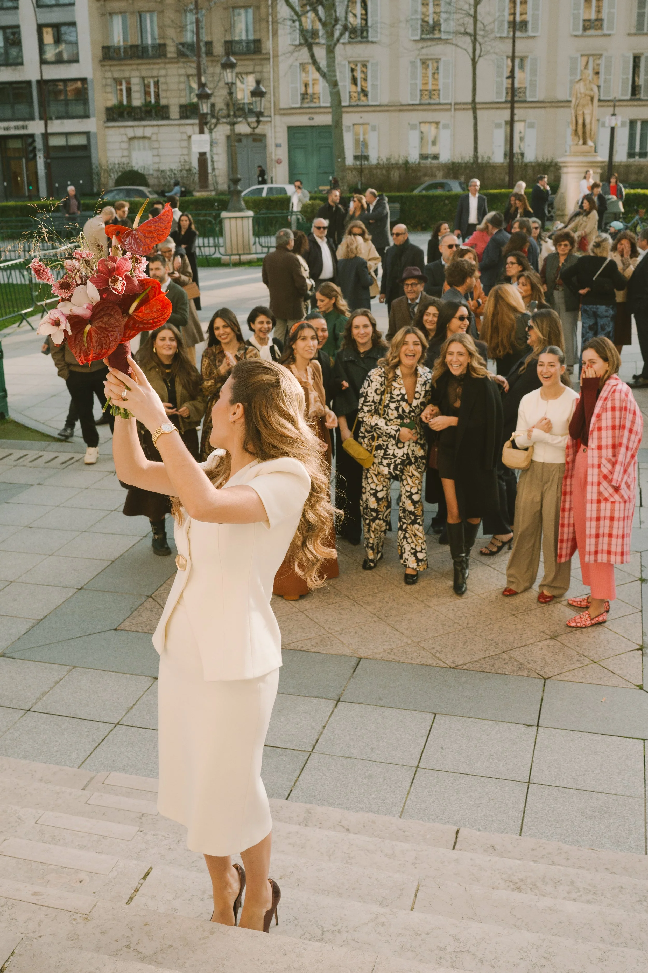 Une femme en robe blanche tient un bouquet de fleurs lors d'une scène de célébration, avec un groupe de personnes souriantes et riantes en arrière-plan dans un espace extérieur urbain.