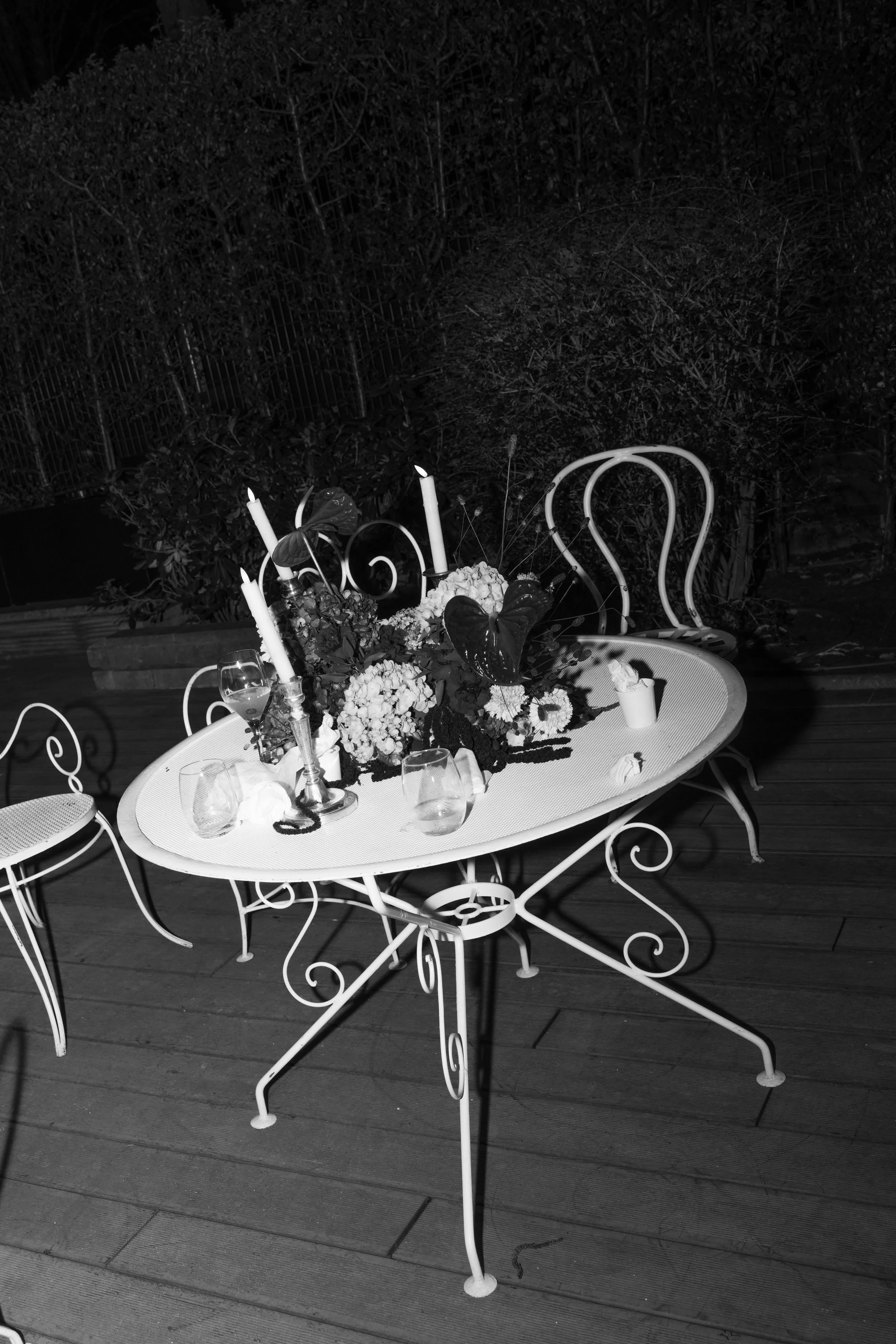 Une table de fer blanc avec des fleurs, des chandeliers, des verres et des bougies dans un jardin nocturne.