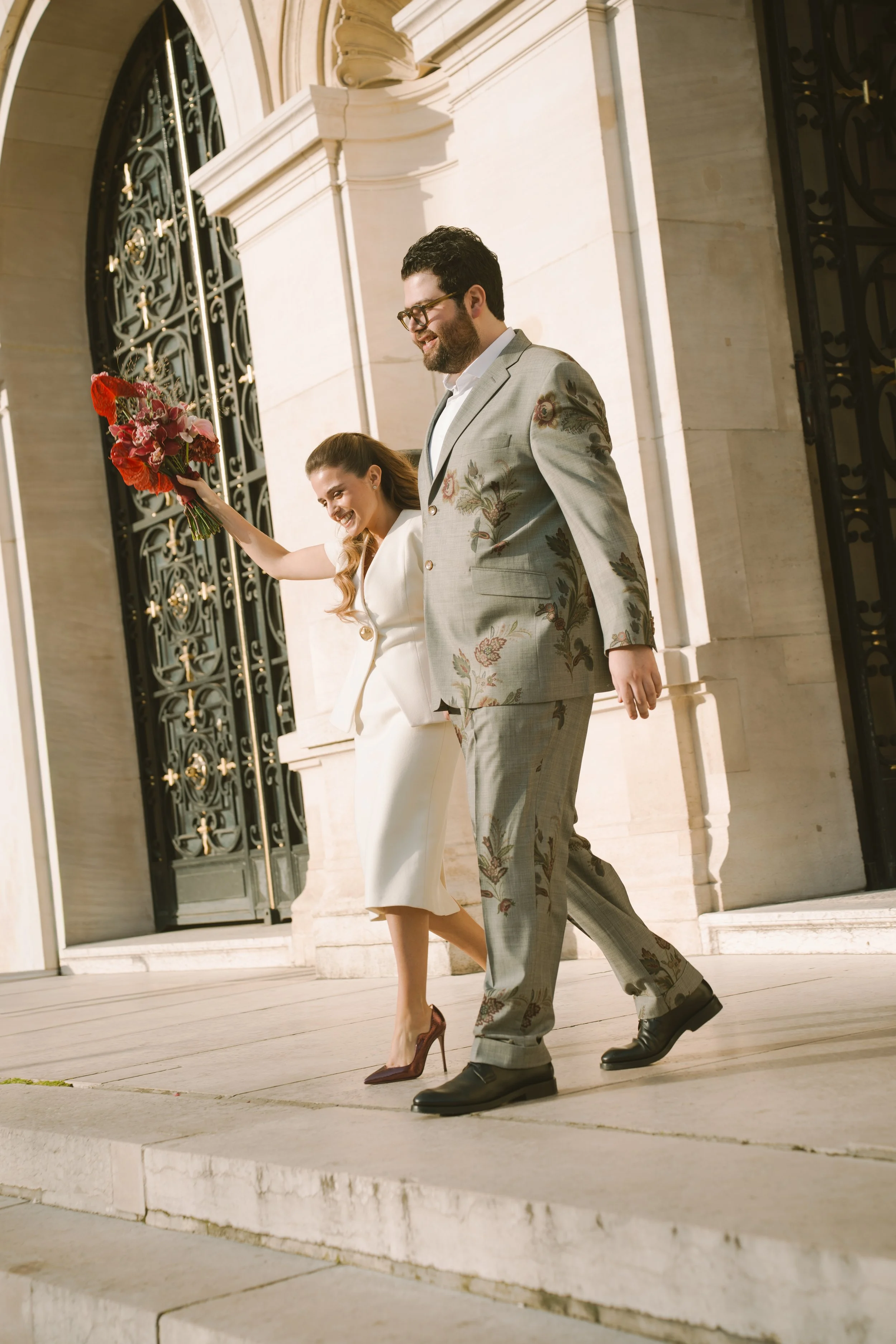 Un couple souriant descend les marches, elle tient un bouquet de fleurs, ils semblent célébrer une occasion spéciale, devant un bâtiment en pierre avec de grandes portes en fer forgé.