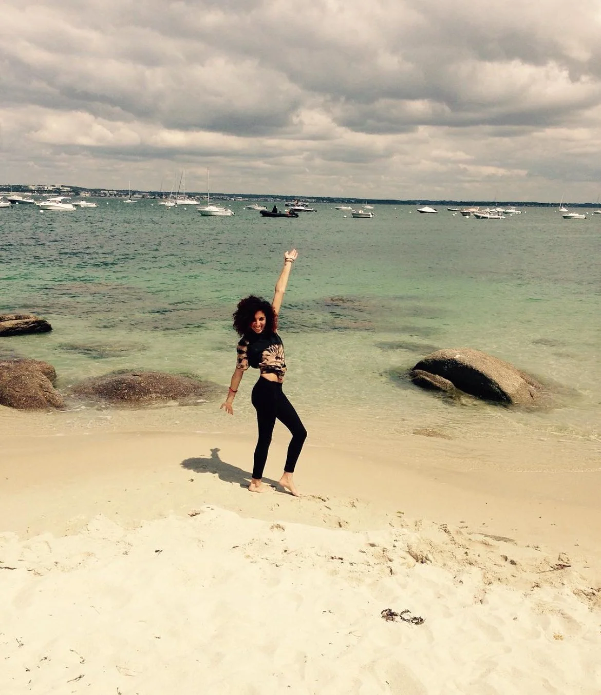 Femme souriante posant sur la plage avec la mer en arrière-plan et des bateaux au loin.