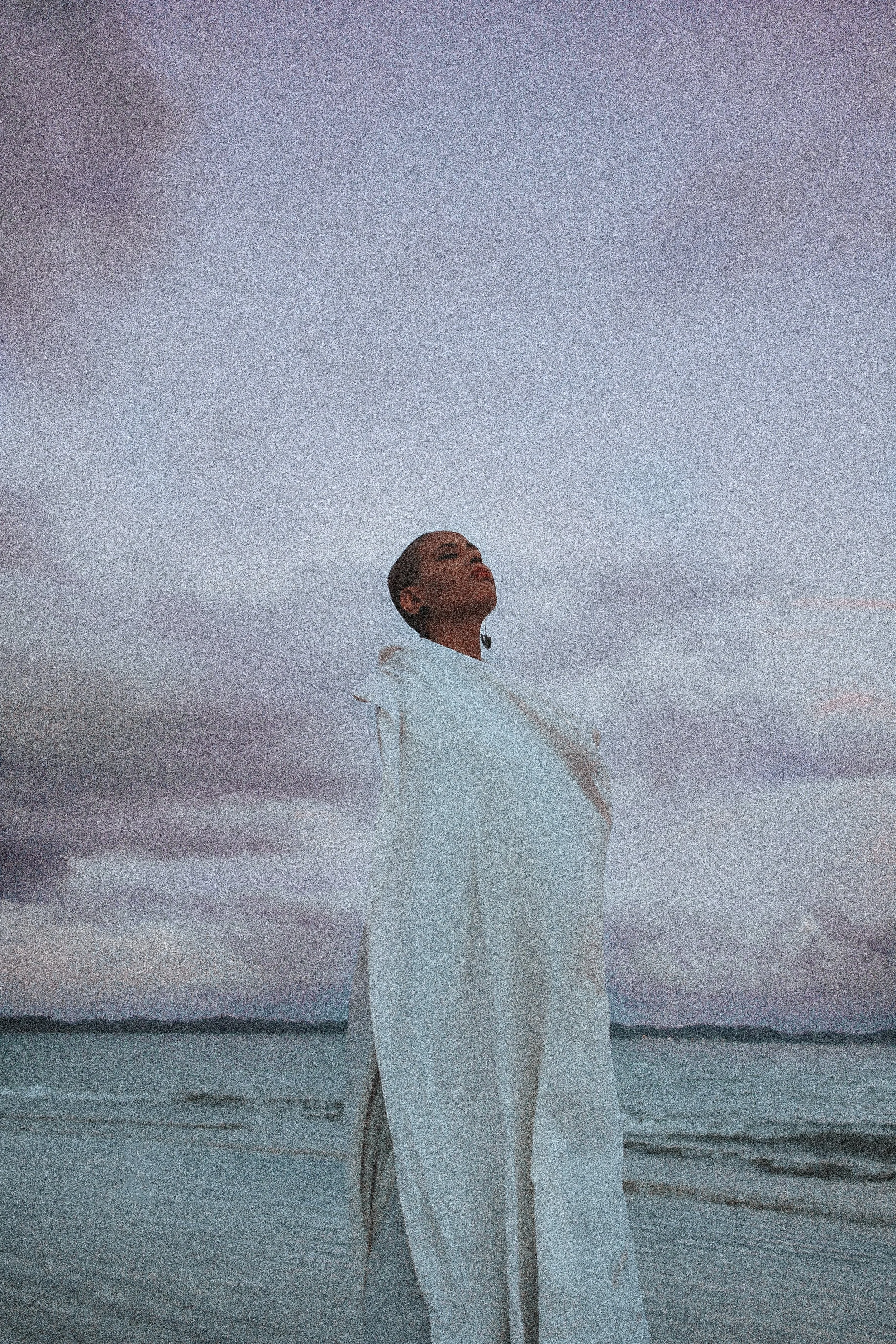 Une femme debout sur la plage avec un ciel nuageux en arrière-plan, portant une robe blanche, regardant vers le haut, dans un contexte de nature et de calme.