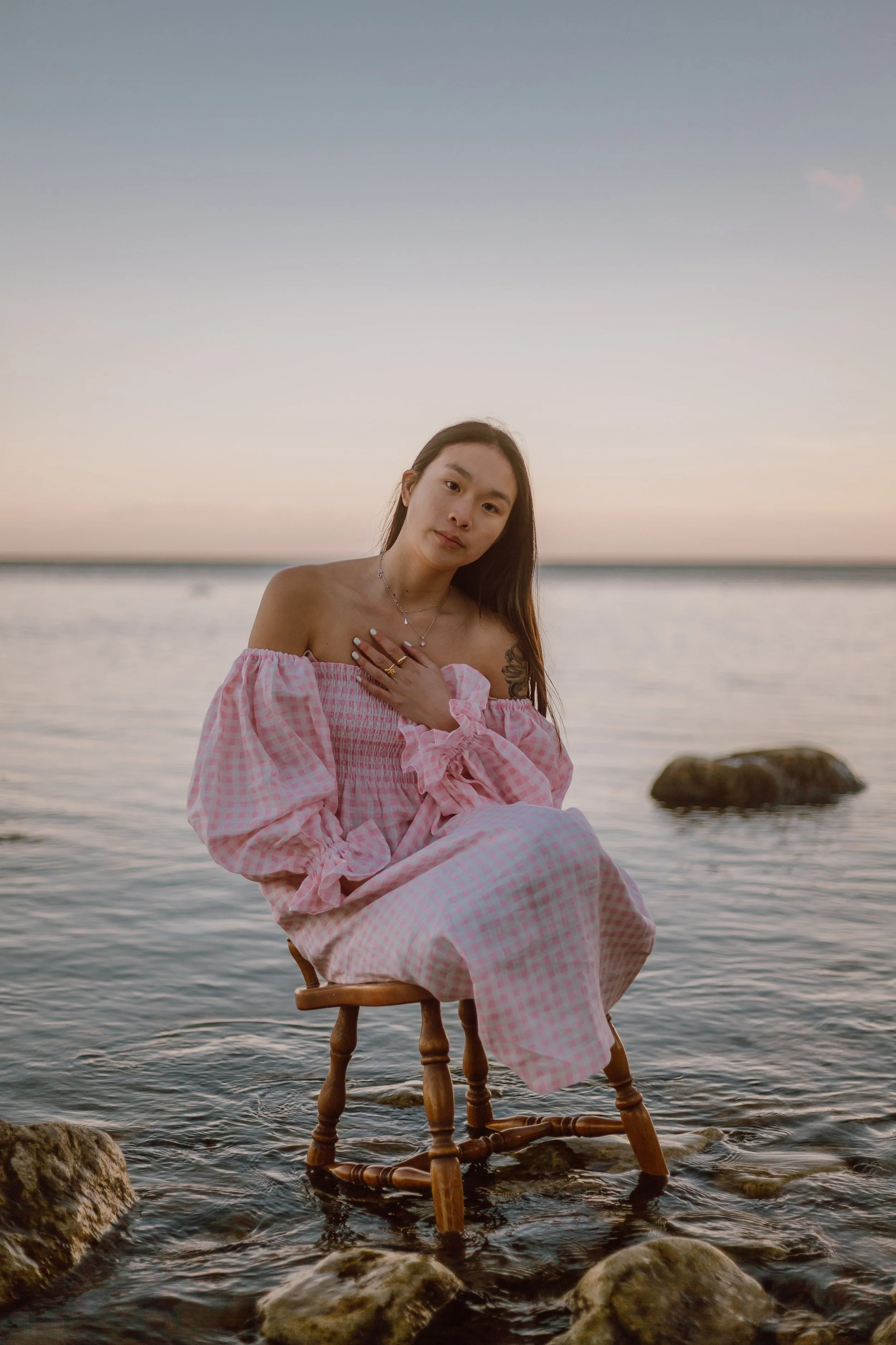 Une femme assise sur une chaise en bois dans l'eau, portant une robe à carreaux rose et blanc, avec un paysage au coucher du soleil en arrière-plan.