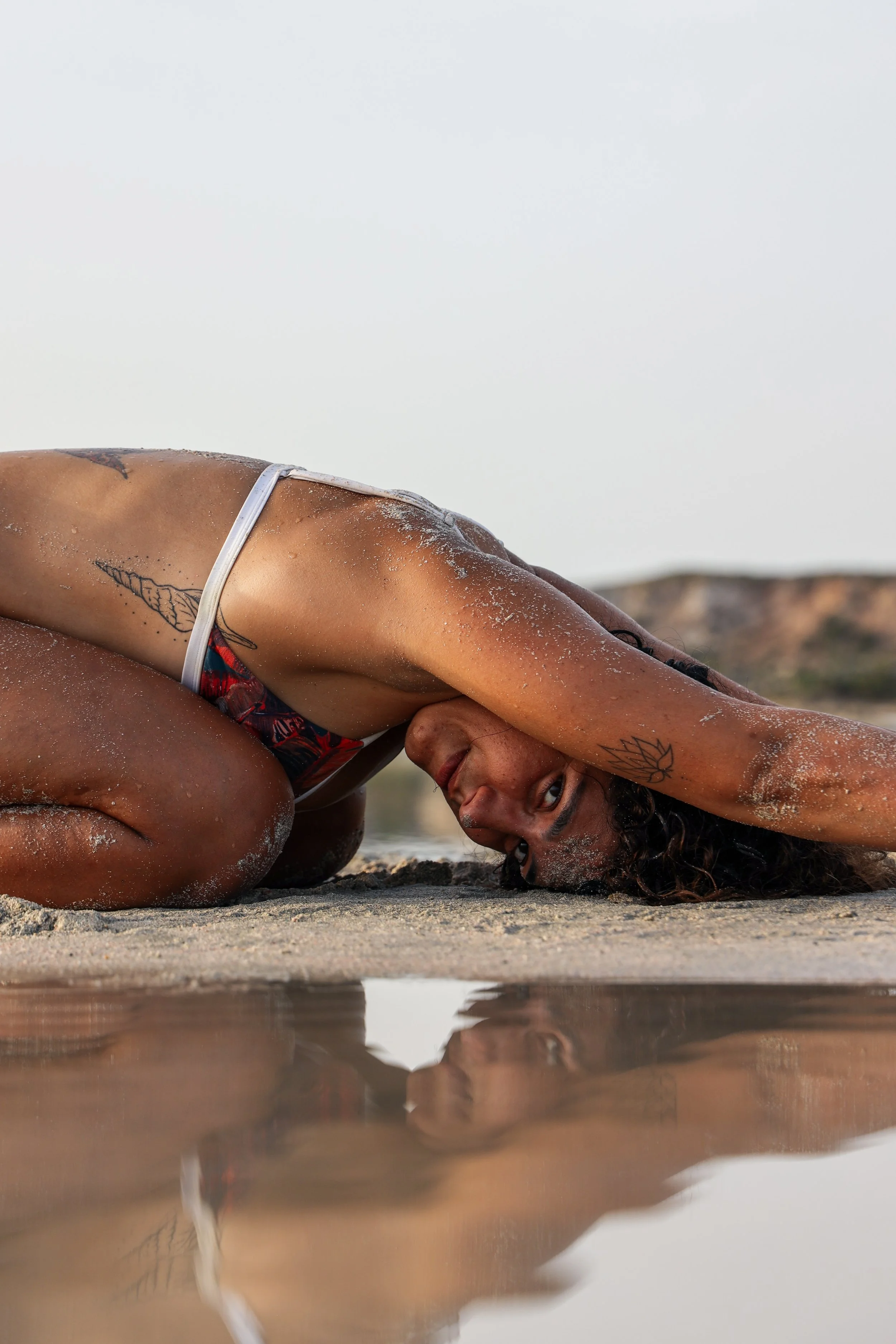 Femme faisant une posture de yoga sur la plage, reflet dans l'eau, paysage côtier au fond.