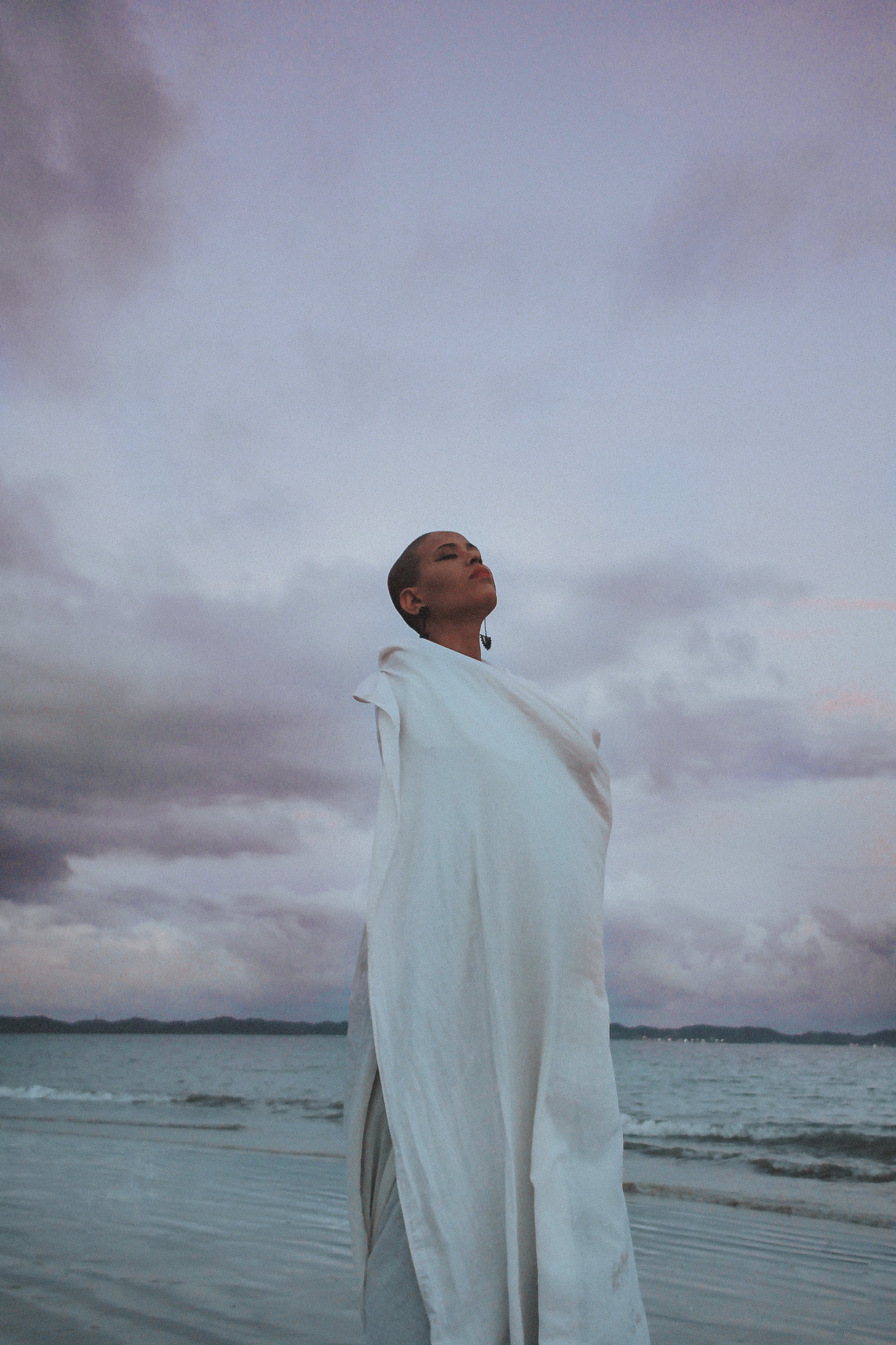 Femme debout sur la plage, portant une robe blanche, regardant vers le ciel avec des nuages sombres en arrière-plan.