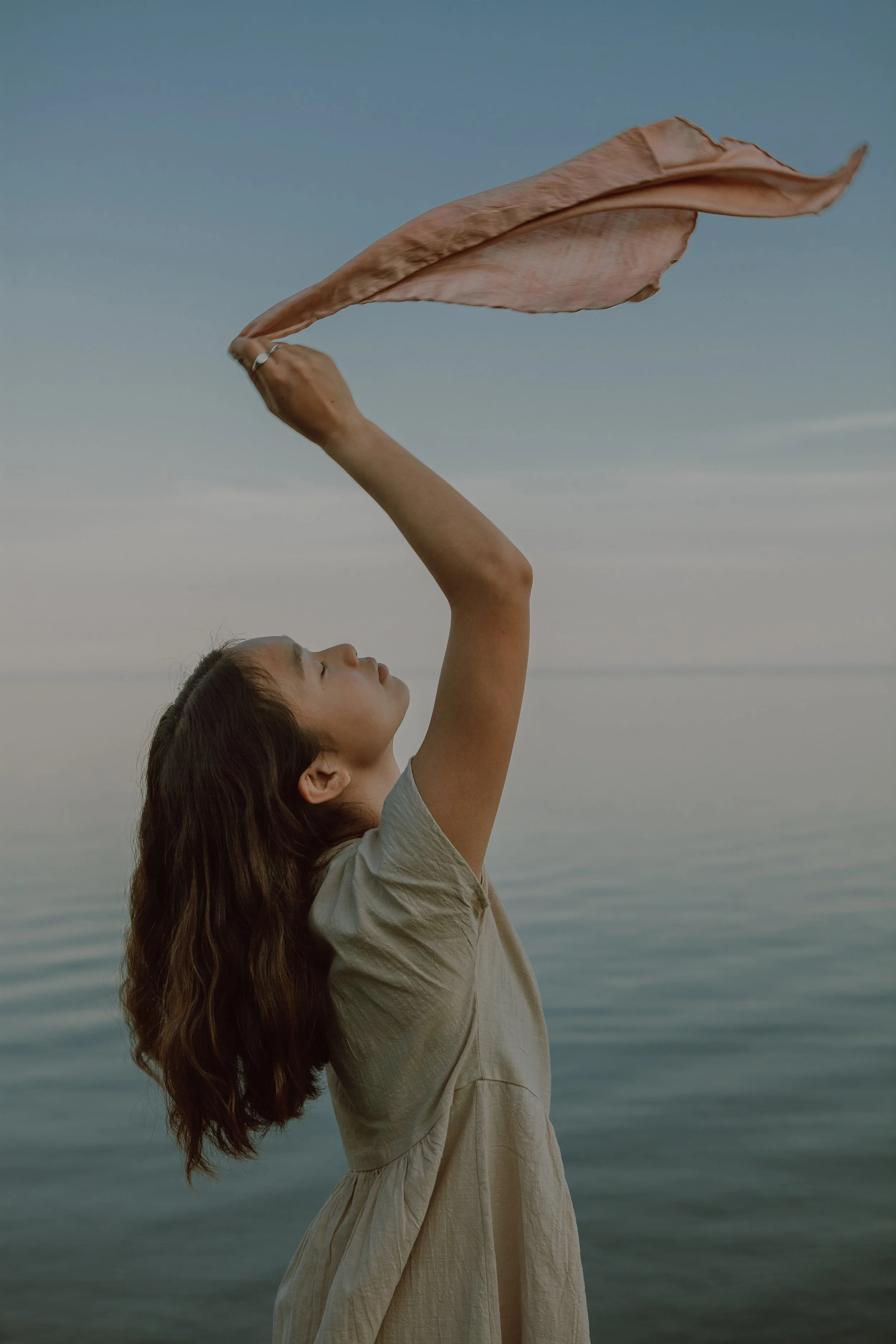 Jeune femme avec cheveux longs, portant une robe beige, levant un foulard en tissu vers le ciel au bord de l'eau, au crépuscule.