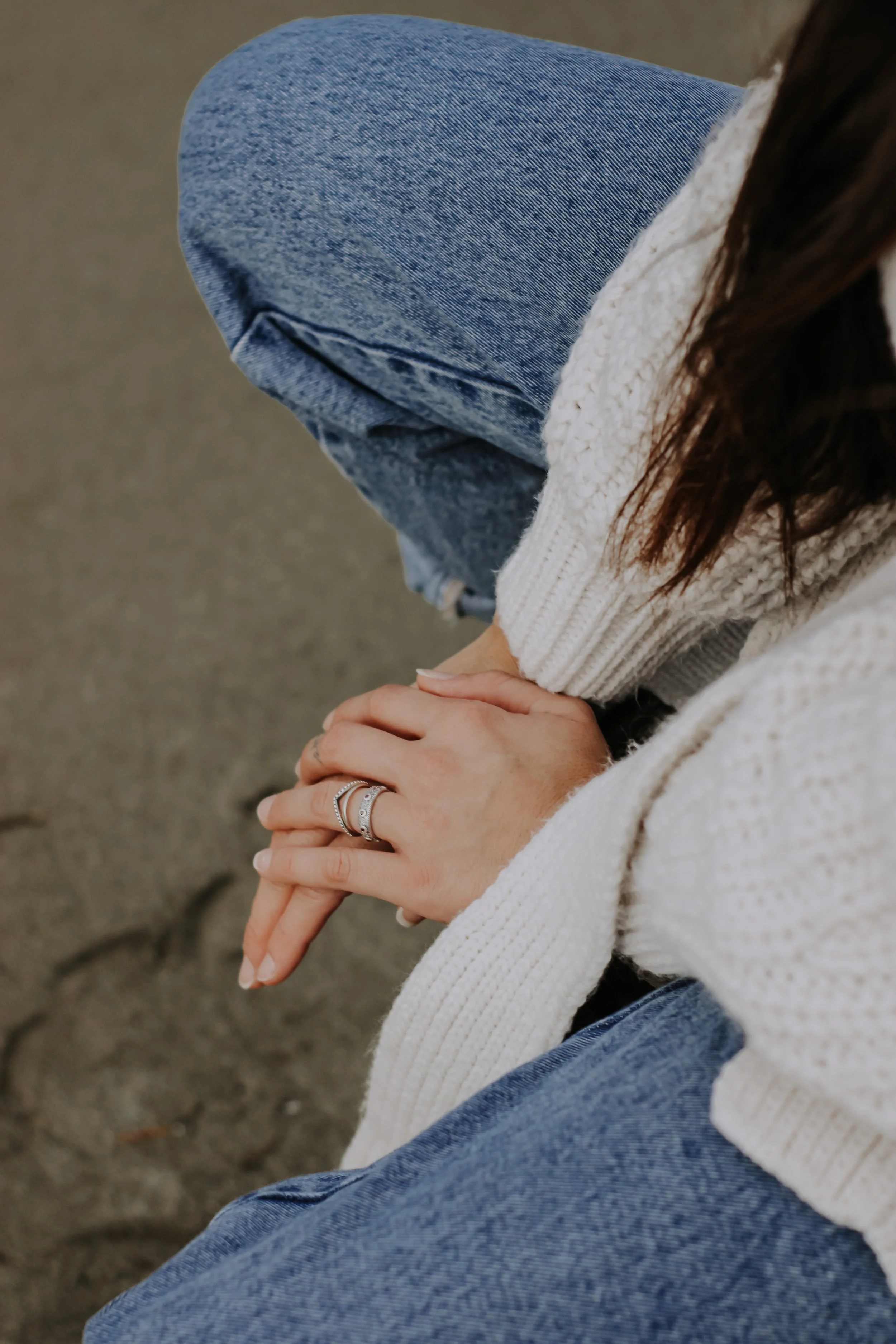 Une femme assise sur du sable, portant un pull blanc, un jean bleu, et plusieurs bagues à la main, avec un aspect détendu et naturel.