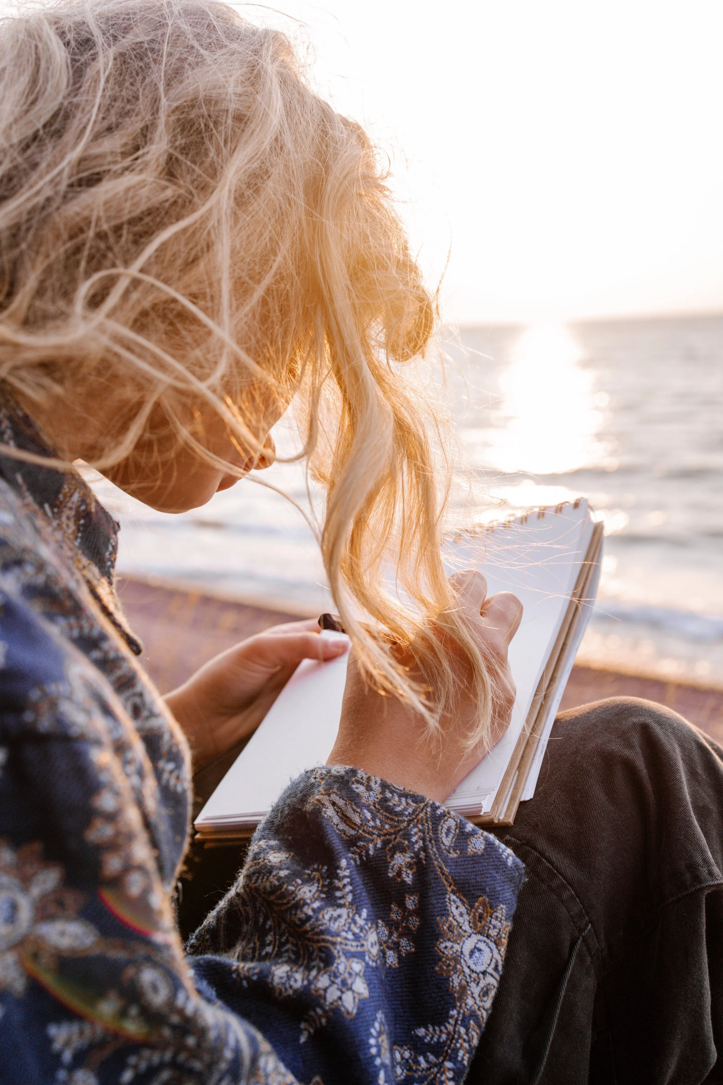 Une femme aux cheveux blonds bouclés écrit dans un carnet au bord de la mer au coucher du soleil.