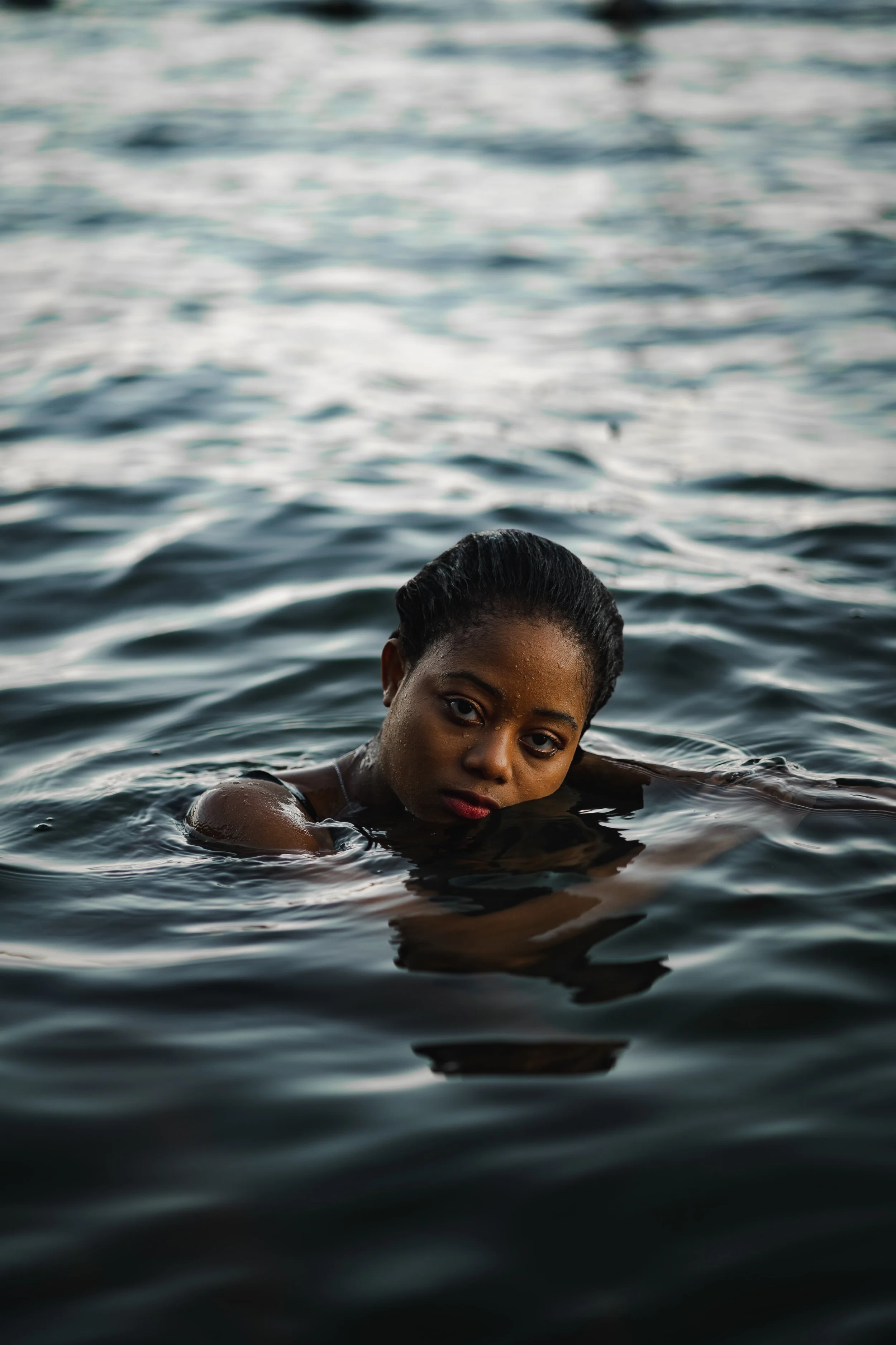 Une femme noire à la peau sombre nage dans l'eau, avec only sa tête et ses épaules visibles, regardant vers l'appareil photo, dans un environnement aquatique calme.