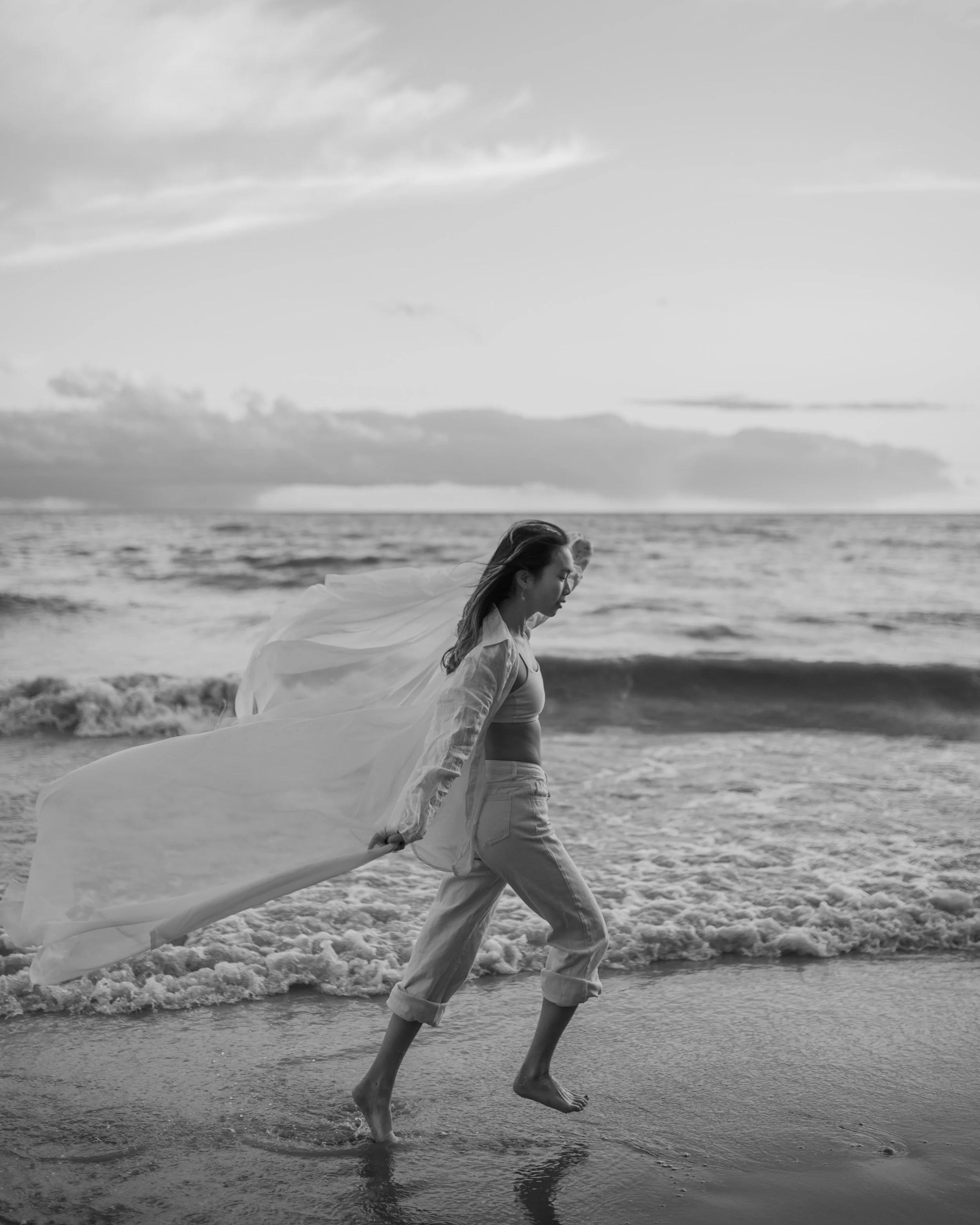 Une femme marche nu-pieds dans l'eau de la mer au coucher du soleil, portant une robe légère et un voile qui souffle dans le vent, en noir et blanc.