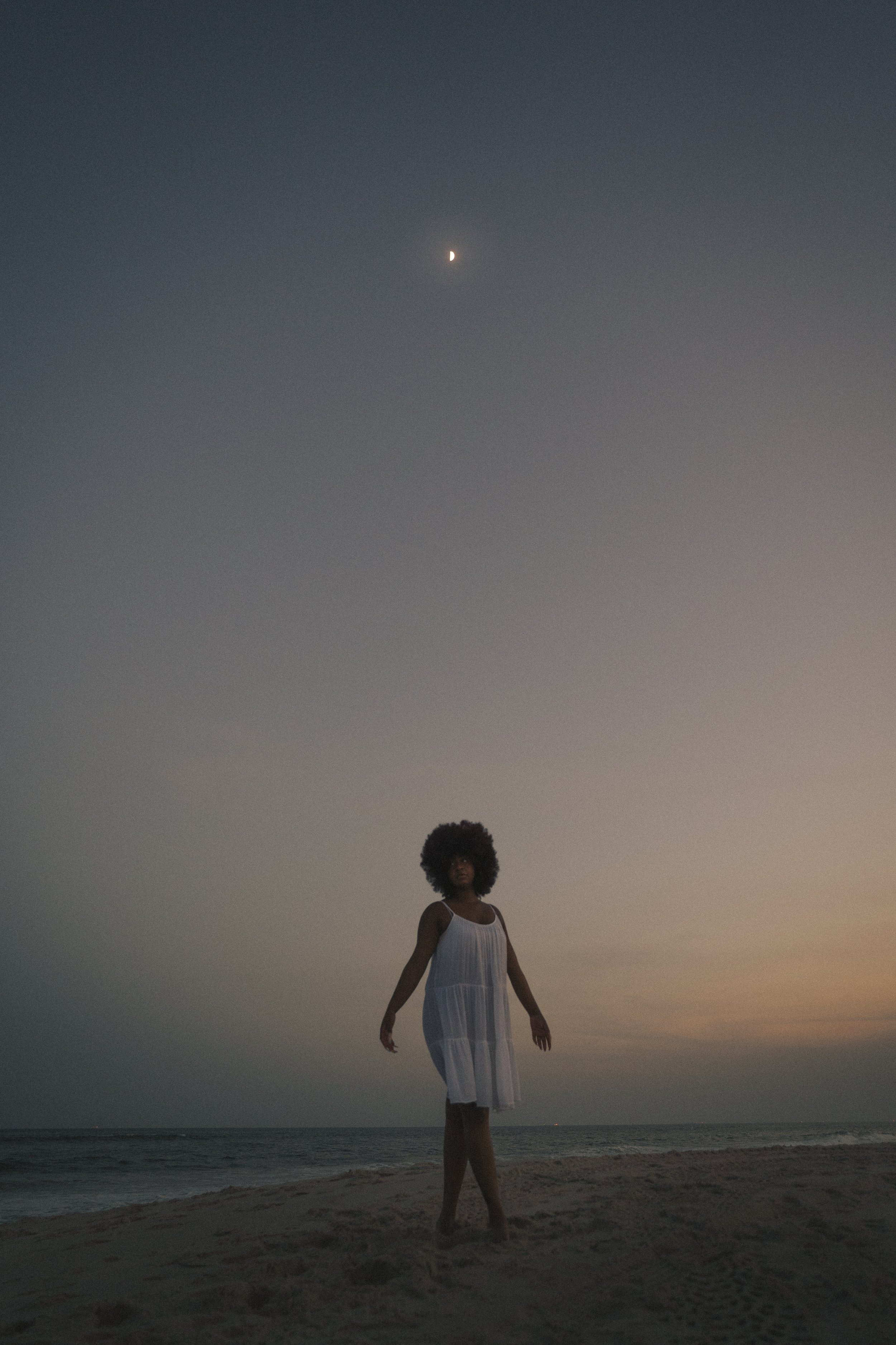 Une femme debout sur la plage au crépuscule, portant une robe blanche, avec la mer en arrière-plan et la lune visible dans le ciel