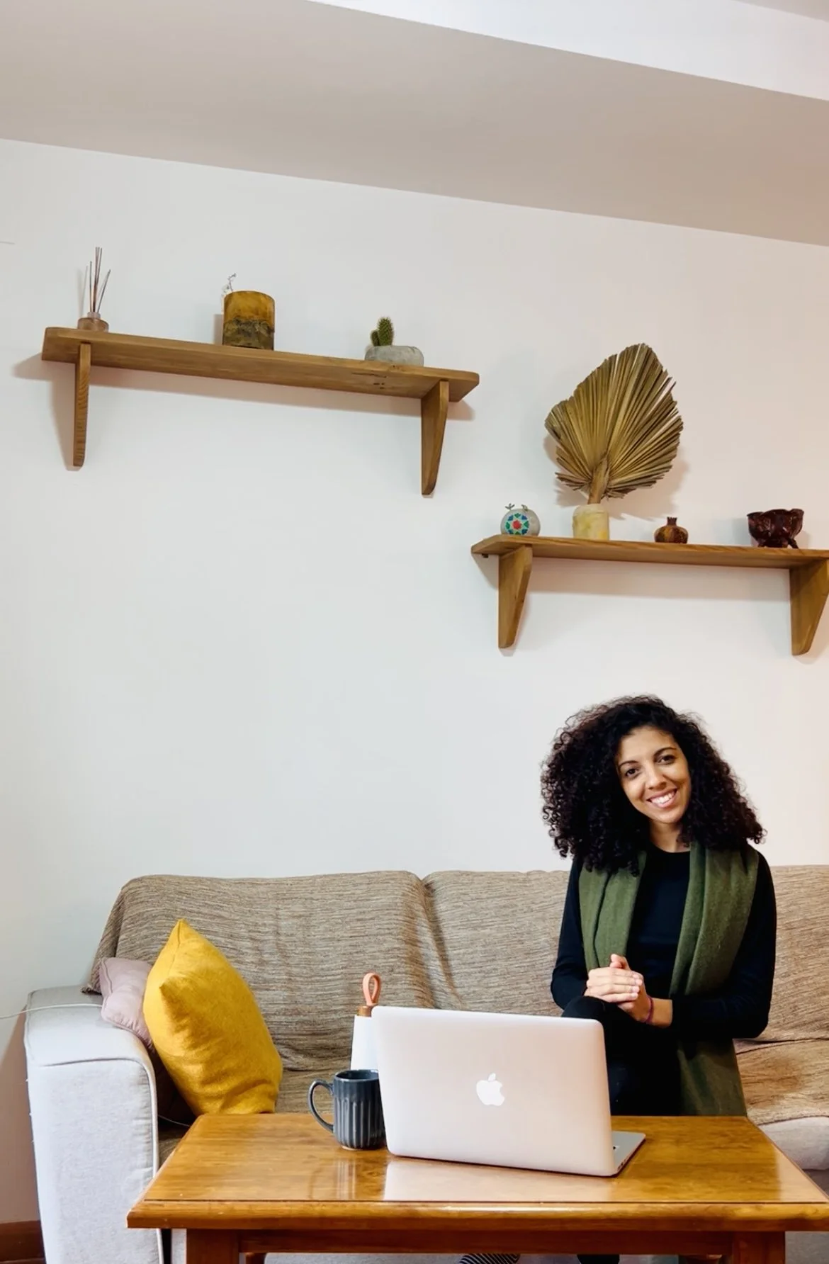 Une femme souriante assise à un bureau en bois avec un ordinateur portable blanc, une tasse noire, dans un salon avec un canapé beige, des coussins jaunes et roses, et deux étagères en bois avec des décorations.