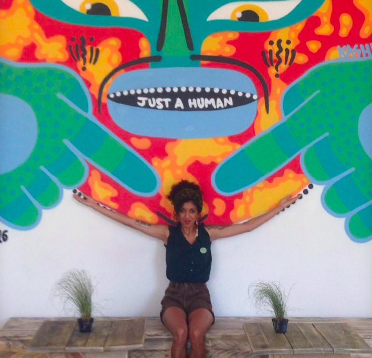 Une femme assise avec les bras étendus devant un mur peint d'un visage coloré avec une bouche grande et un message "JUST A HUMAN" dans la bouche.