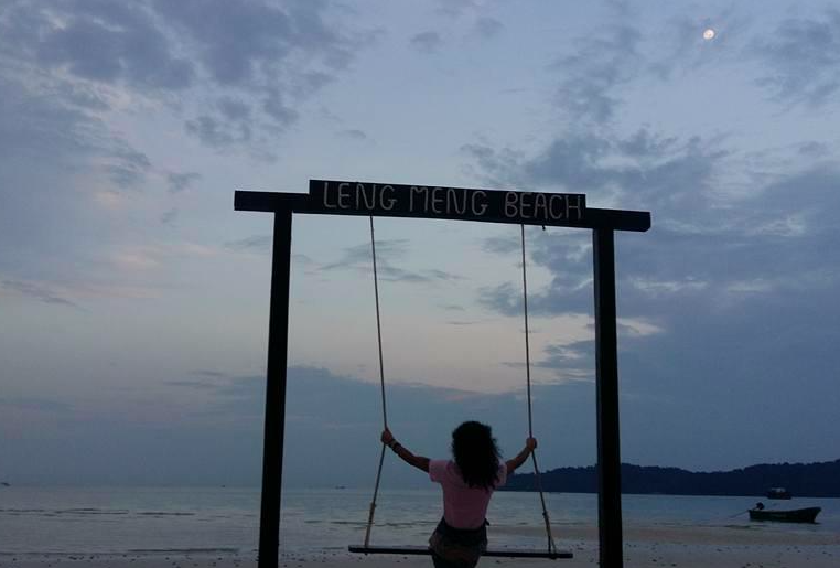 Une femme assise sur une balançoire en bord de mer à LENG MENG BEACH, avec un ciel nuageux et la lune visible.