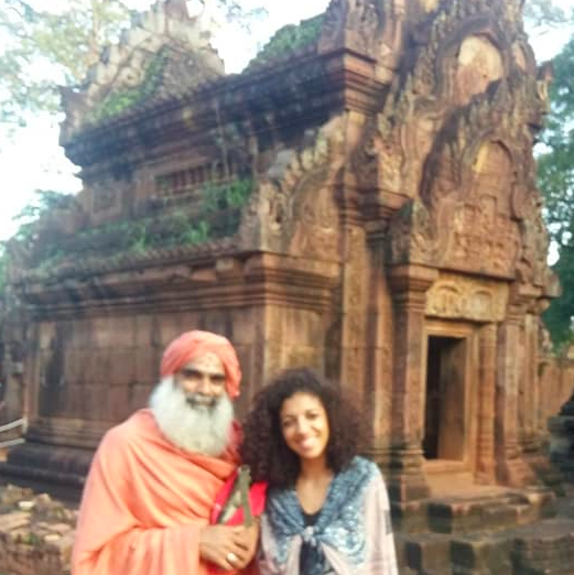 Un homme âgé en vêtements traditionnels et une jeune femme souriante posent devant un ancien monument en pierre rouge, entouré de verdure.
