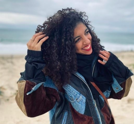 Femme souriante avec cheveux bouclés, portant une veste colorée, sur la plage par temps nuageux.