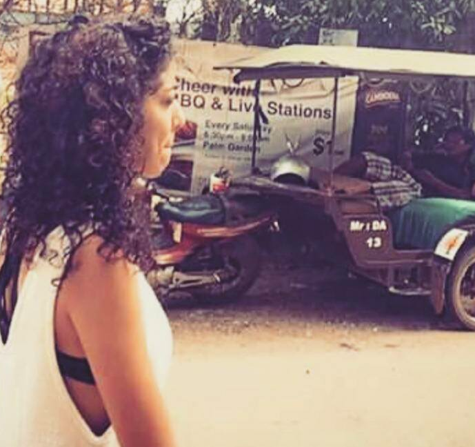 Une femme à cheveux bouclés regarde vers la gauche, avec une station de barbecue et nourriture en arrière-plan, notamment un chariot et une voiturette dans un environnement en plein air.