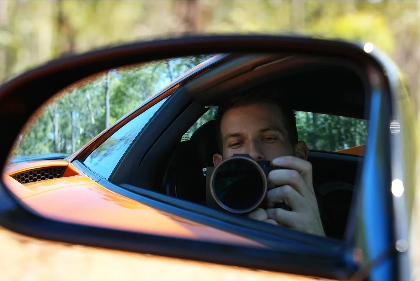 A man taking a selfie with a camera in a car's side mirror, with green trees in the background.