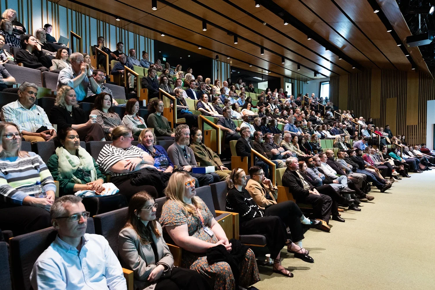 Audience sitting in theater-style seats attending a conference or presentation in a modern auditorium with wooden ceiling panels and vertical wall accents.
