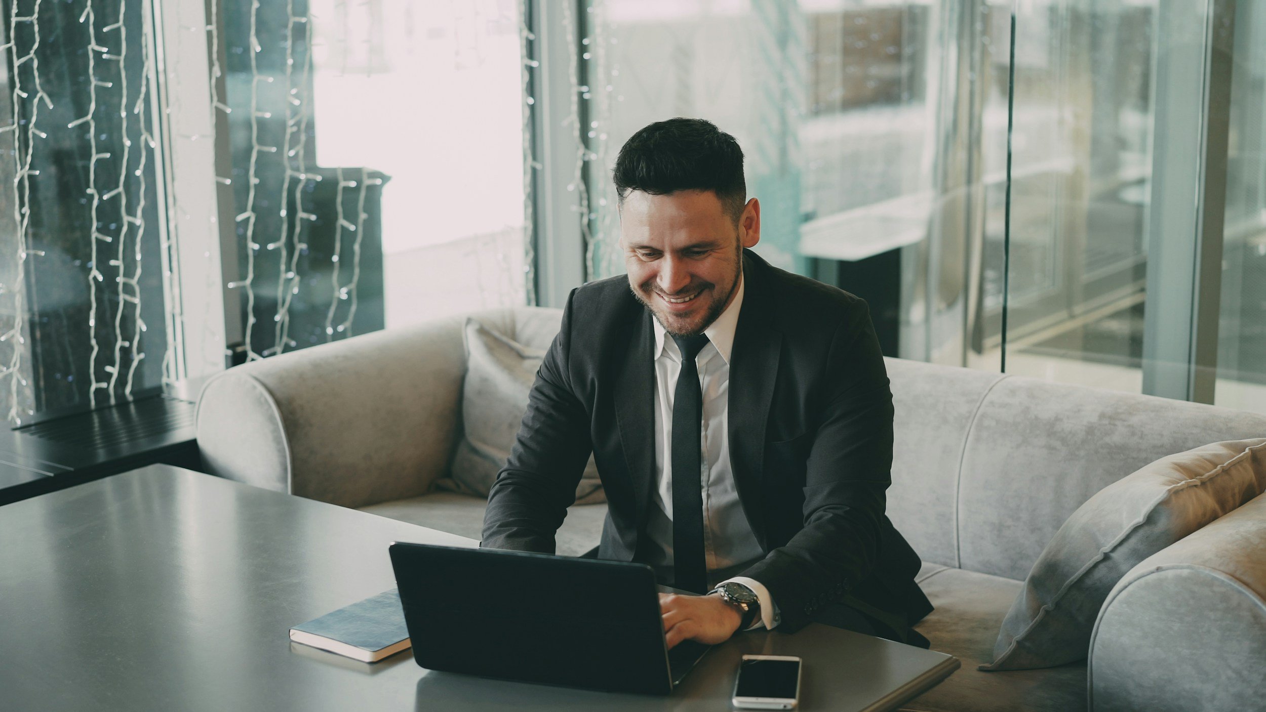 A man in a black suit and tie sitting on a sofa working on a laptop in a modern office space with large glass windows and decorations.