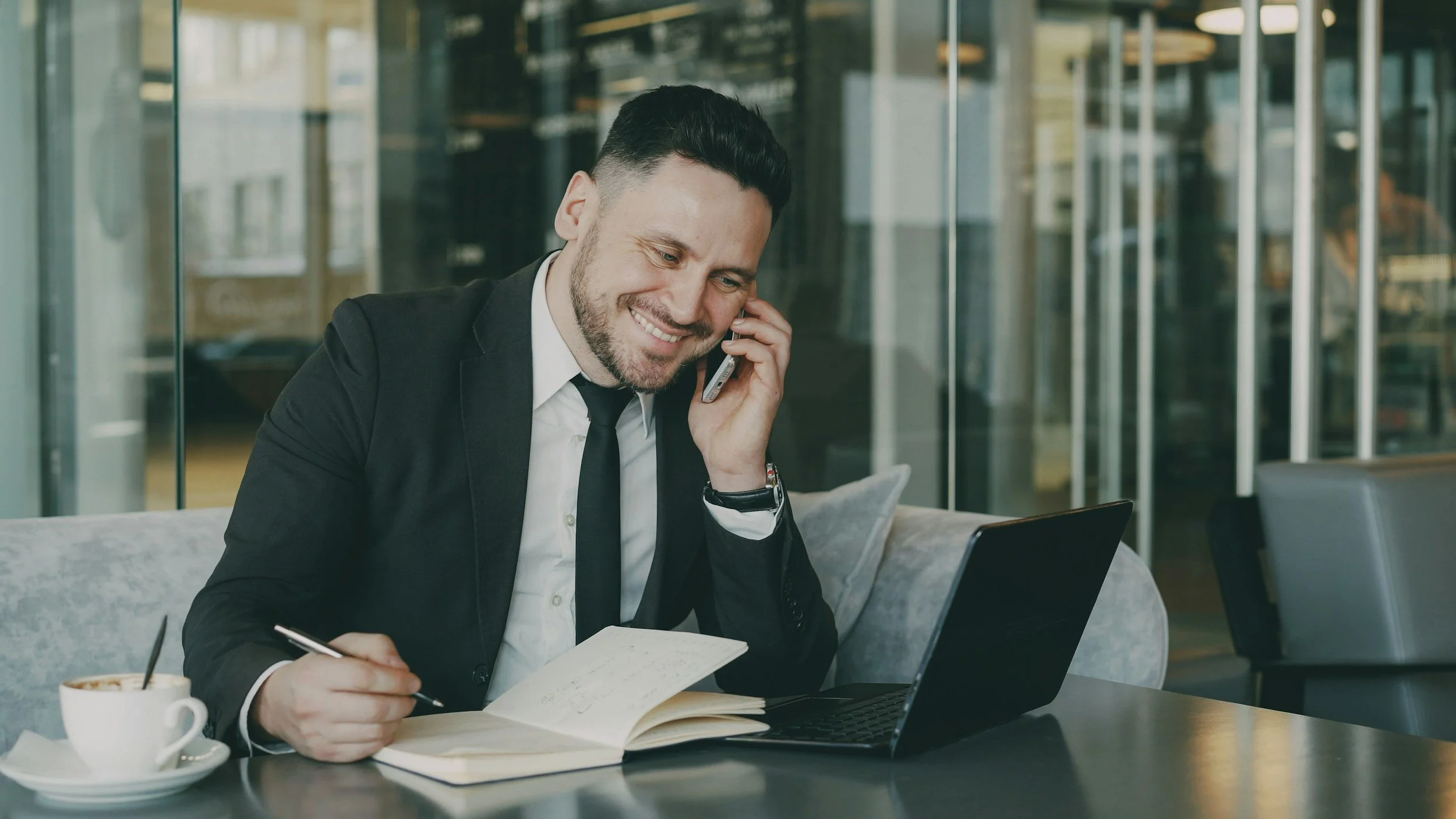 Businessman in a black suit and white shirt, sitting at a table, talking on a mobile phone, smiling, with a laptop, notebook, pen, and coffee in a modern office setting.