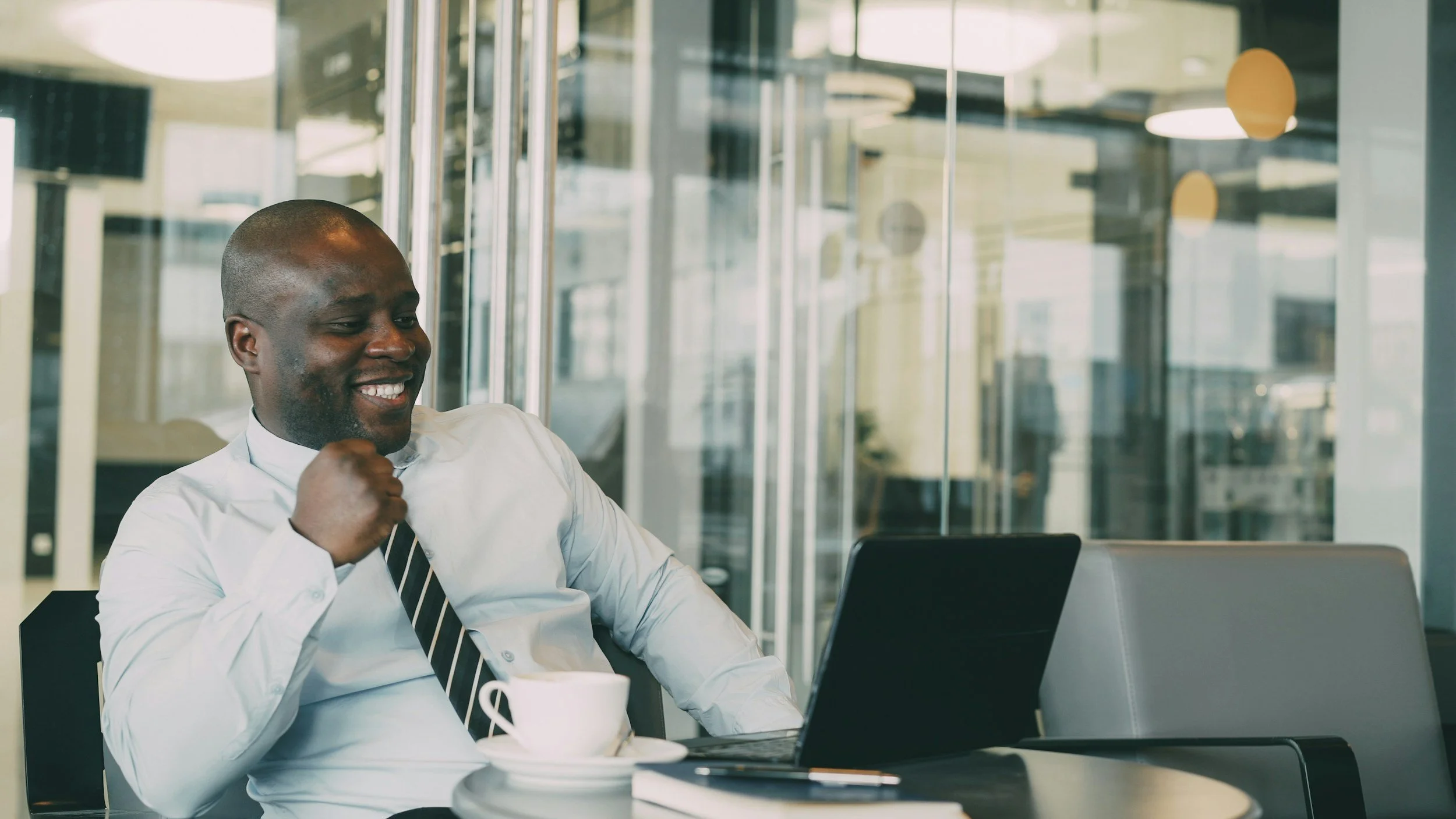 A smiling man in a white shirt and striped tie sitting at a table in an office or cafe, working on a laptop with a coffee cup and a notebook in front of him.