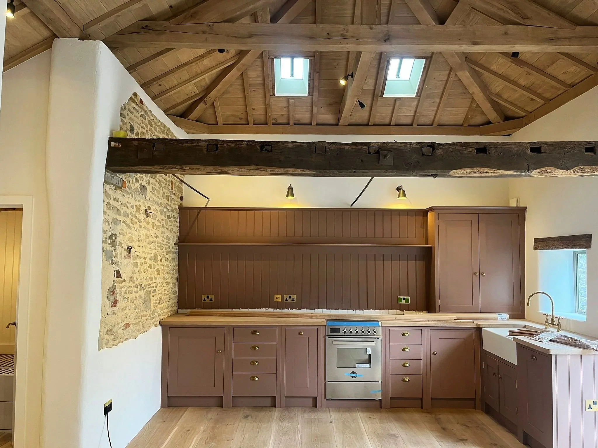 Interior view of a kitchen with pink cabinetry, a wood countertop, an oven, and a window. The ceiling is wooden with skylights, and there is a rustic wooden beam running across the room. An exposed stone wall partial is visible on the left.
