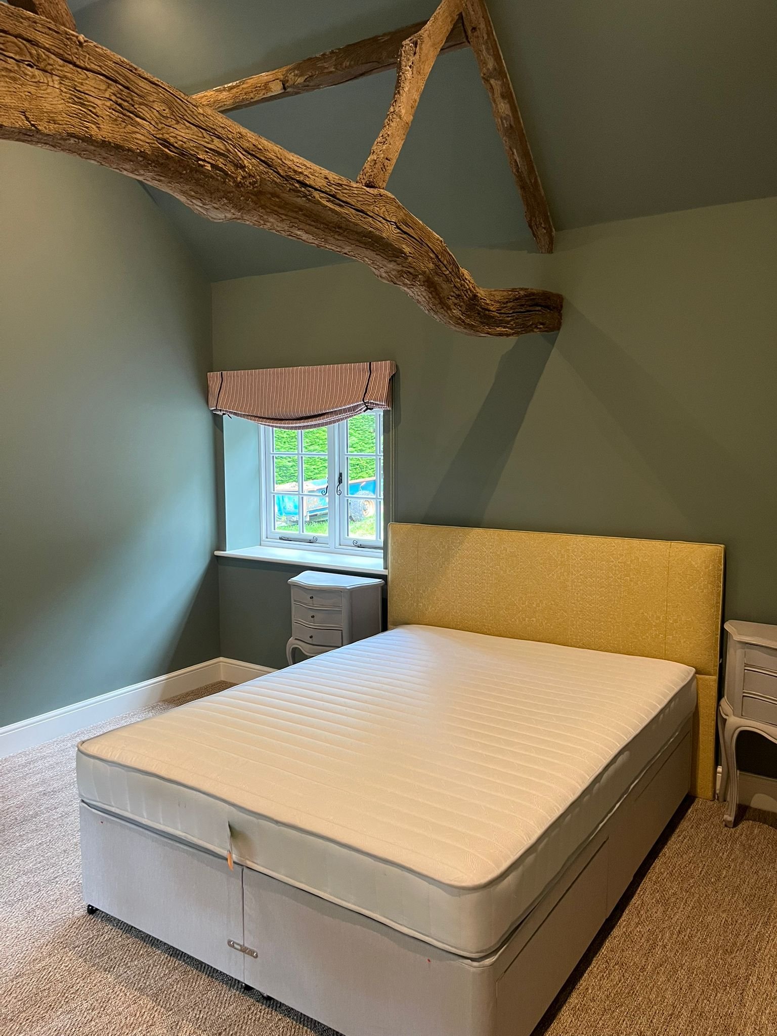 A bedroom with a yellow headboard, a white mattress, two small nightstands, a window with striped valance, and wooden beams on the ceiling.