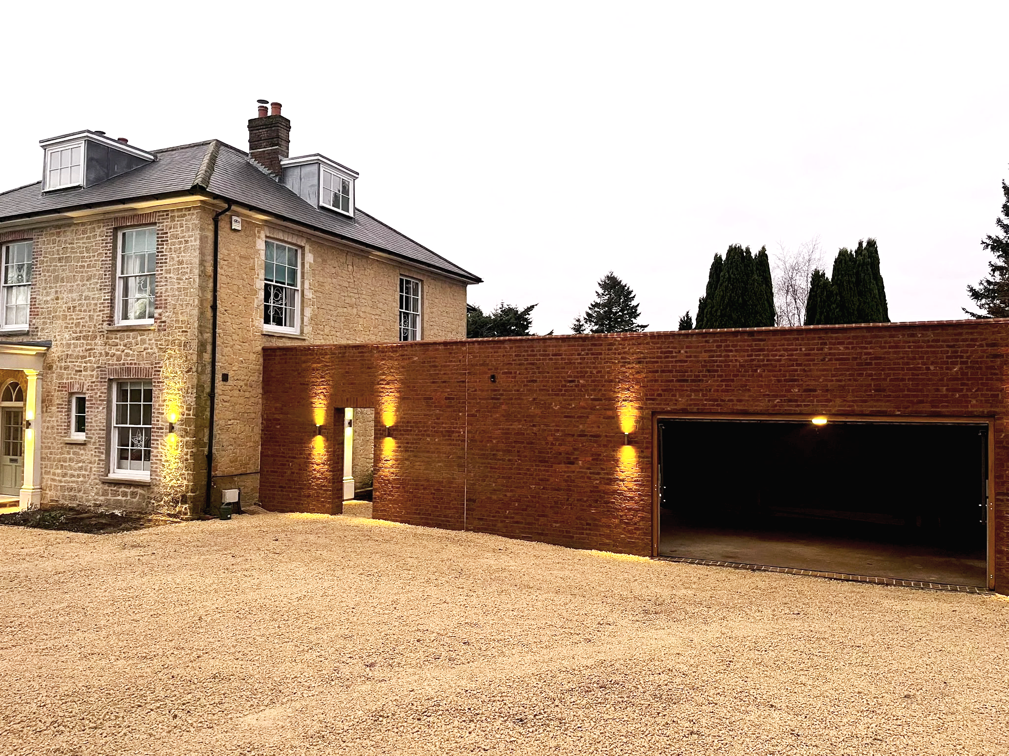 A large brick house with white-framed windows and a gray roof, with a modern brick garage attached. The garage has outdoor lighting illuminating the brick wall, and the driveway is covered with gravel. Tall trees are visible in the background against an overcast sky.